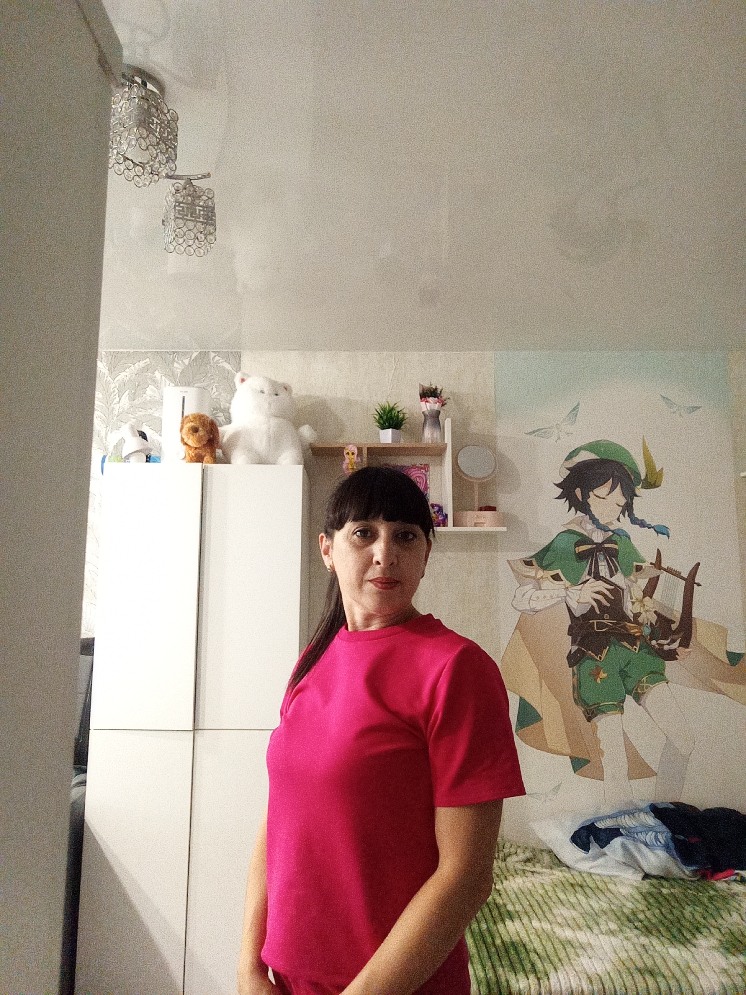The image shows a medium shot of a woman with dark hair and bangs, wearing a bright pink shirt, standing in a room. She is facing the camera with a neutral expression. Behind her, to the left, is a white cabinet with decorative items on top, including a white teddy bear and a small brown stuffed dog. To the right of the cabinet is a wall with a large anime-style mural of a character holding a lyre. Above the cabinet, a chrome light fixture with crystal accents hangs from the ceiling. The room appears to be a bedroom, with a bed covered in a green and white striped blanket visible in the lower right corner. The overall lighting is artificial and somewhat dim, with a slight grainy quality to the image. The location is identified as Belogorsk, Russia.