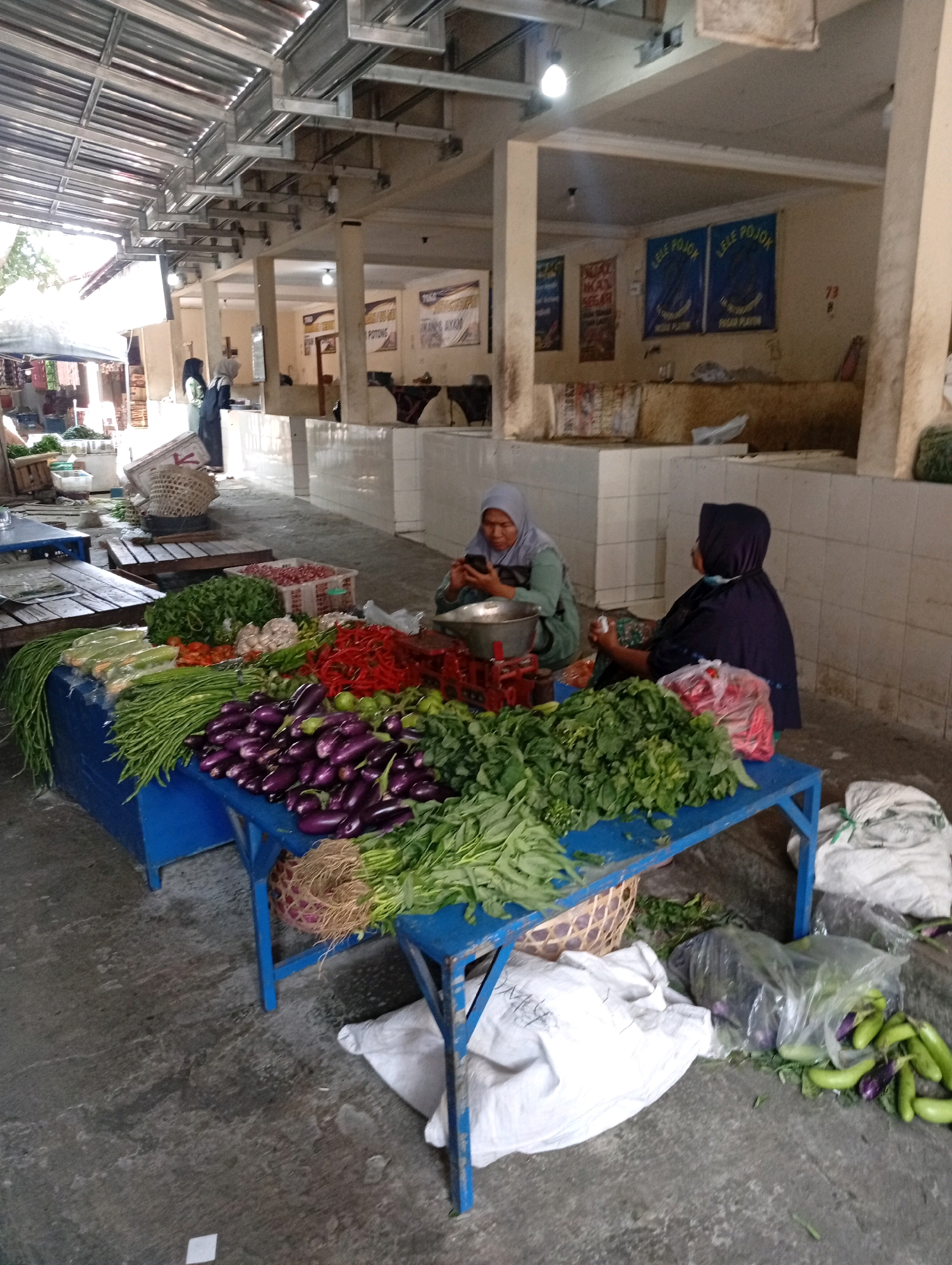 The image depicts a vibrant outdoor market scene in Playen, Indonesia. The foreground is dominated by a blue table laden with an abundant array of fresh produce. Varieties of green vegetables, including long beans and leafy greens, are piled high, alongside bright red chilies, eggplants, and other root vegetables. Behind the produce, two women are visible. One woman, wearing a light blue hijab, is seated and appears to be operating a weighing scale. The other woman, in a dark hijab, is also seated, with her back partially turned to the camera. The market is set against a backdrop of stalls and structures, suggesting a bustling commercial environment. The ground is a concrete surface, typical of market pathways. Several woven baskets and bags are scattered around, adding to the market's authenticity. The overall impression is one of a lively marketplace showcasing local agricultural products and the daily activities of vendors in Playen.