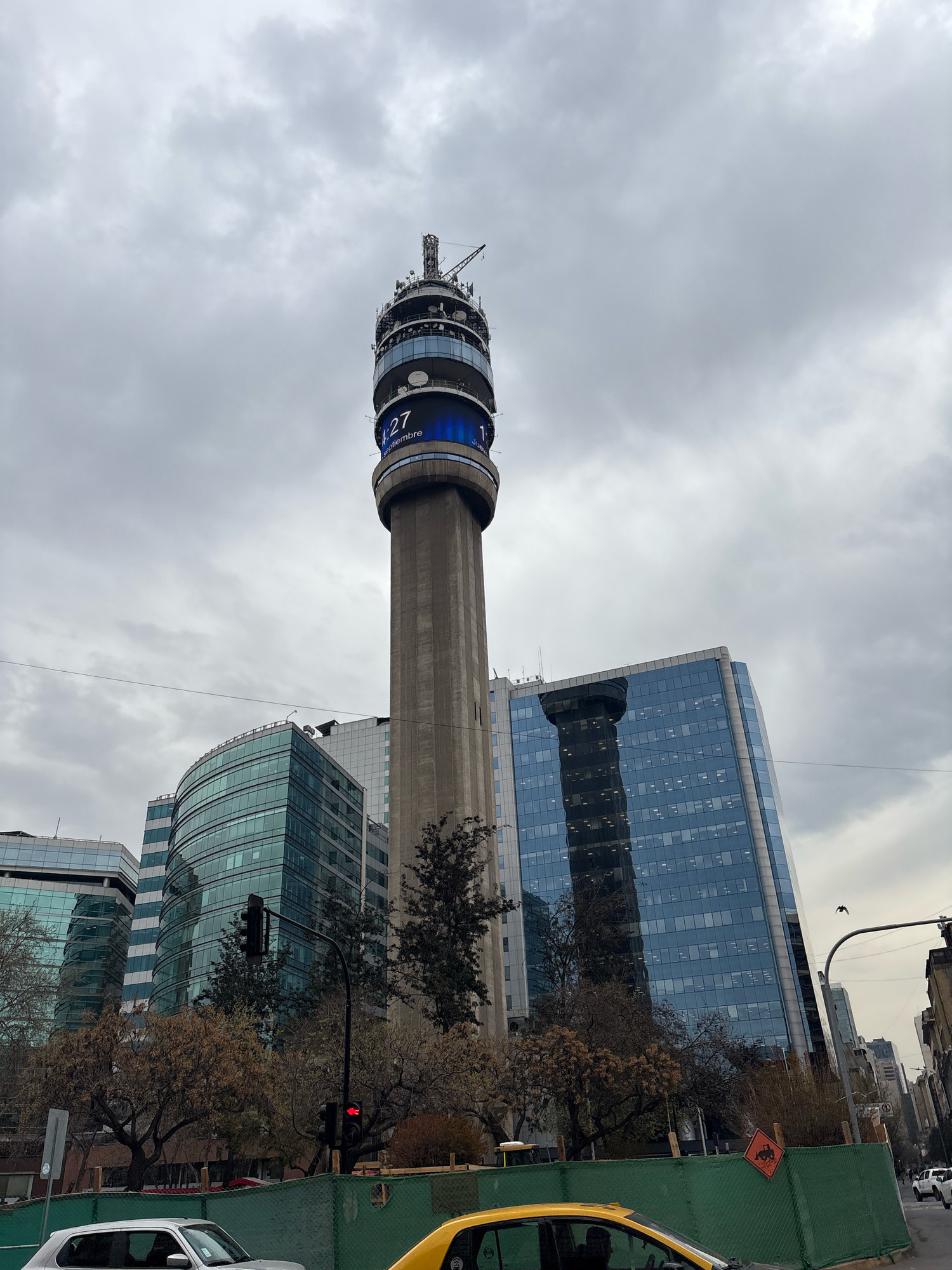 The image shows a view of the Costanera Center Tower in Santiago, Chile, on an overcast day. The tower, a prominent landmark, dominates the center of the frame. It is a tall, concrete structure with a circular observation deck at the top, featuring a large digital display showing "27" and "11" with Spanish text below. Surrounding the tower are modern, glass-faced buildings, some of which reflect the sky and the tower itself. In the foreground, there is a street scene with a yellow taxi in the lower right corner, and a silver car to the left. Traffic lights are visible, one of which is red. A green construction fence runs across the bottom of the image, with an orange construction sign depicting a bulldozer. Bare trees with brown foliage are scattered throughout the scene, suggesting it may be autumn or winter. The sky is filled with thick, grey clouds. A bird is captured in flight against the cloudy sky.