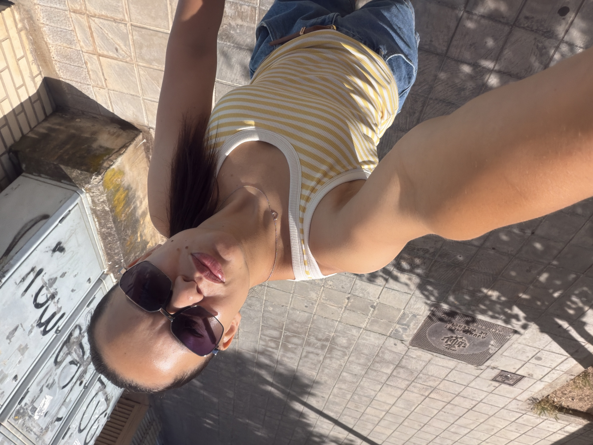 A high-angle, medium shot captures a woman from the chest up, posing for a selfie outdoors on a sunny day. She is wearing a white and yellow striped tank top and denim shorts. Large, dark-lensed sunglasses with metallic frames obscure her eyes. Her dark brown hair is pulled back into a ponytail. She has on a delicate silver necklace. The background shows a tiled sidewalk, a metal grate embedded in the pavement with an emblem that reads "AYUNTAMENT DE VALENCIA," and a graffitied white structure on the right side of the frame. Shadows from unseen trees or structures are cast across the ground.