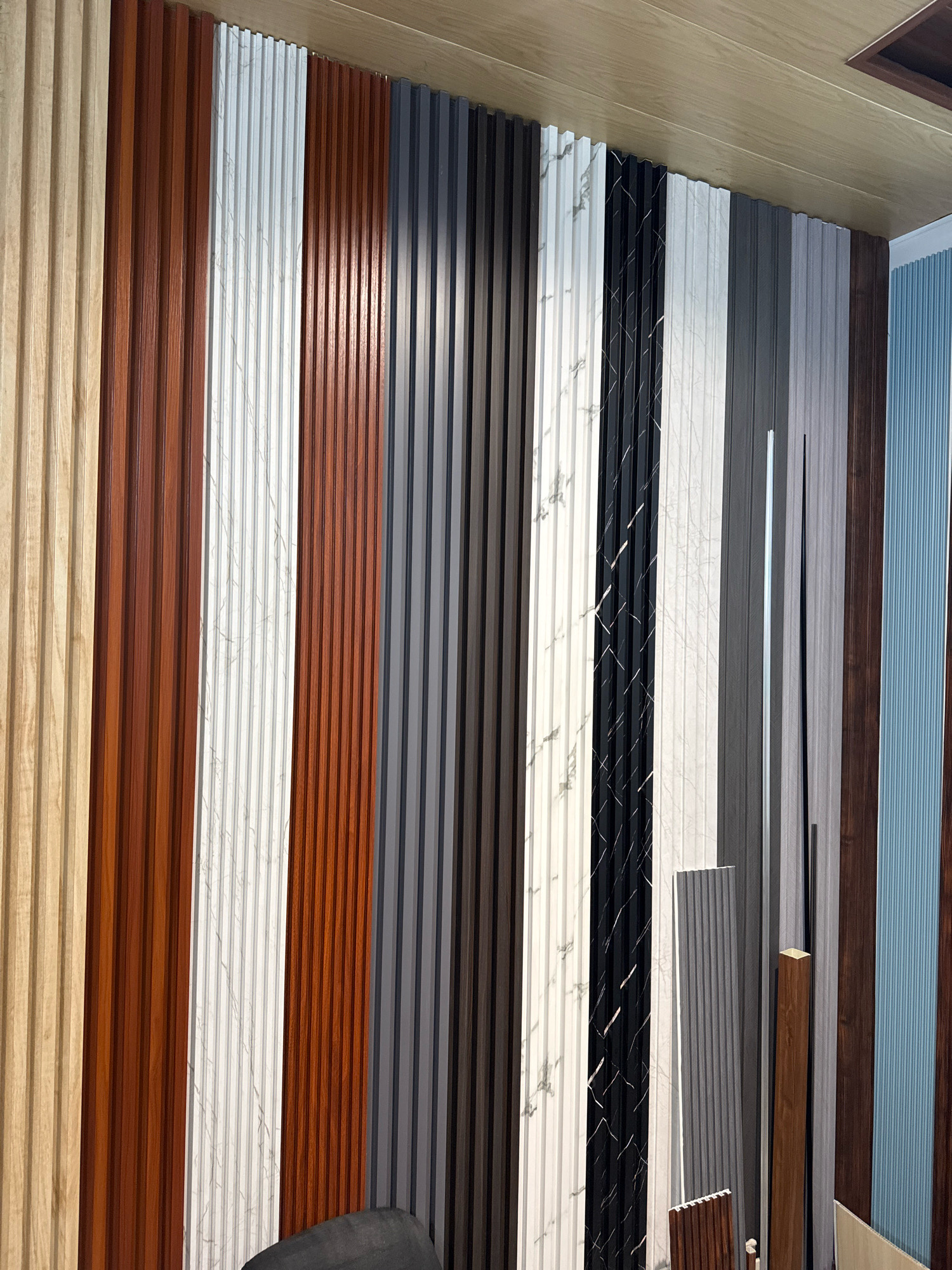 The image shows a wall covered with various decorative panels, likely in a showroom or a construction site in Jagadhari, India. The panels are arranged vertically and exhibit a range of textures and colors, including light wood grain, dark wood grain, white marble-like patterns, black marble-like patterns, and solid gray and dark gray colors. Some panels have a ribbed or grooved texture, while others appear smoother. In the foreground, a dark gray chair is partially visible at the bottom center. There are also some thin, vertical metal or plastic pieces and a wooden accent piece leaning against the wall. The ceiling above appears to be made of light-colored wooden panels. The overall scene depicts a display of interior design materials.