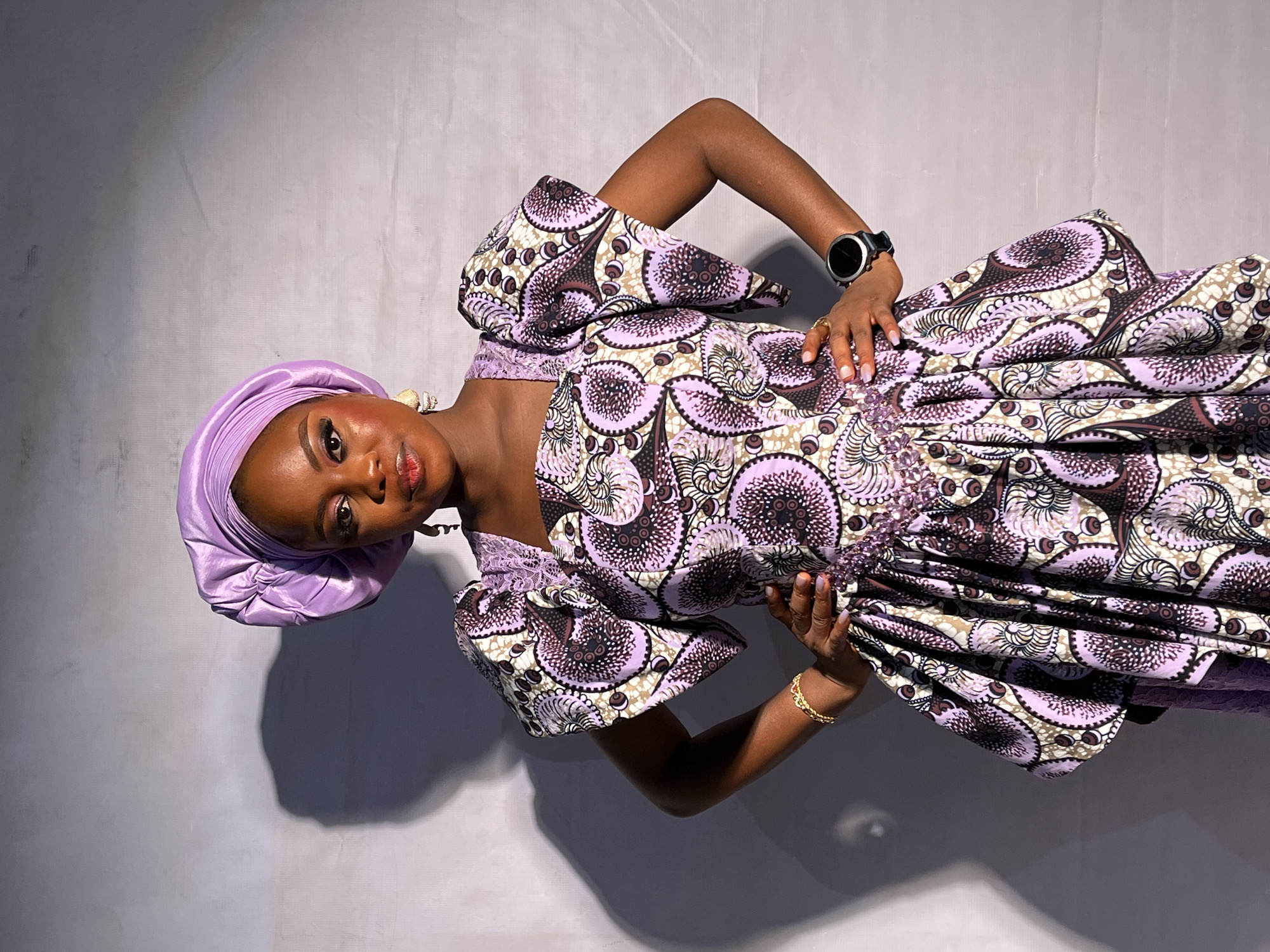 A young woman stands indoors against a light gray or white wall. She wears a traditional lavender-purple patterned dress with large sleeves and a matching headwrap. She has dark skin, with a watch on her left wrist and a gold bracelet on her right wrist. Her hands are placed on her waist and lower torso. Her expression is neutral as she looks directly at the viewer. The background is out of focus. The location is Barakin Akawo, Nigeria.
