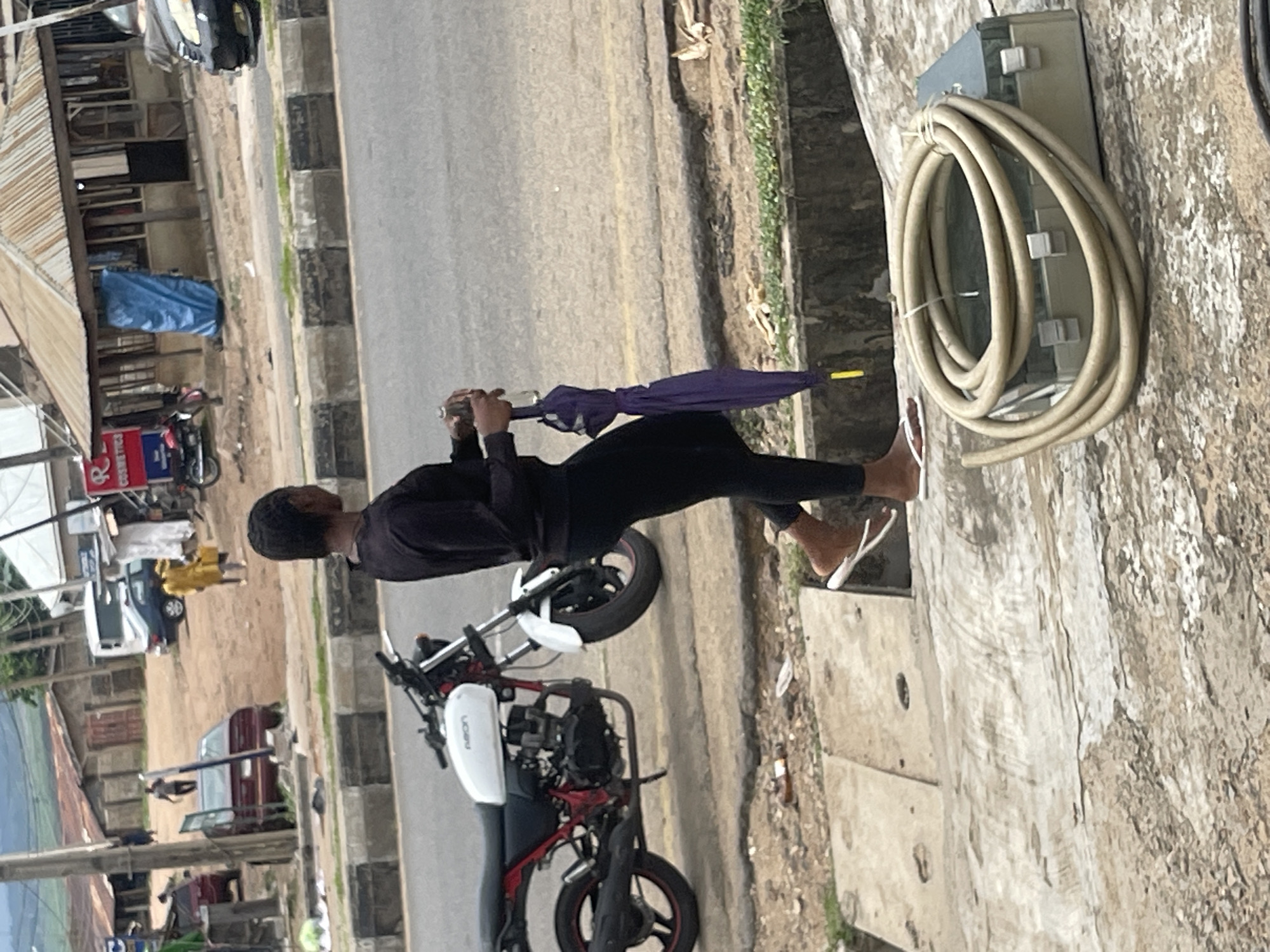 A woman, seen from the back, walks on a paved surface near a road. She is wearing a black long-sleeved shirt and black leggings, with white flip-flops on her feet. In her right hand, she holds a clear bottle, and in her left hand, she carries a folded purple umbrella with a yellow tip. A white motorcycle with red accents is parked to her left. In the background, there are buildings, some with corrugated metal roofs, and several vehicles. The ground in the foreground has scattered debris, including a coiled hose on top of a light-colored box. The environment appears to be an urban or semi-urban area, possibly in a developing country given the architecture and road conditions.