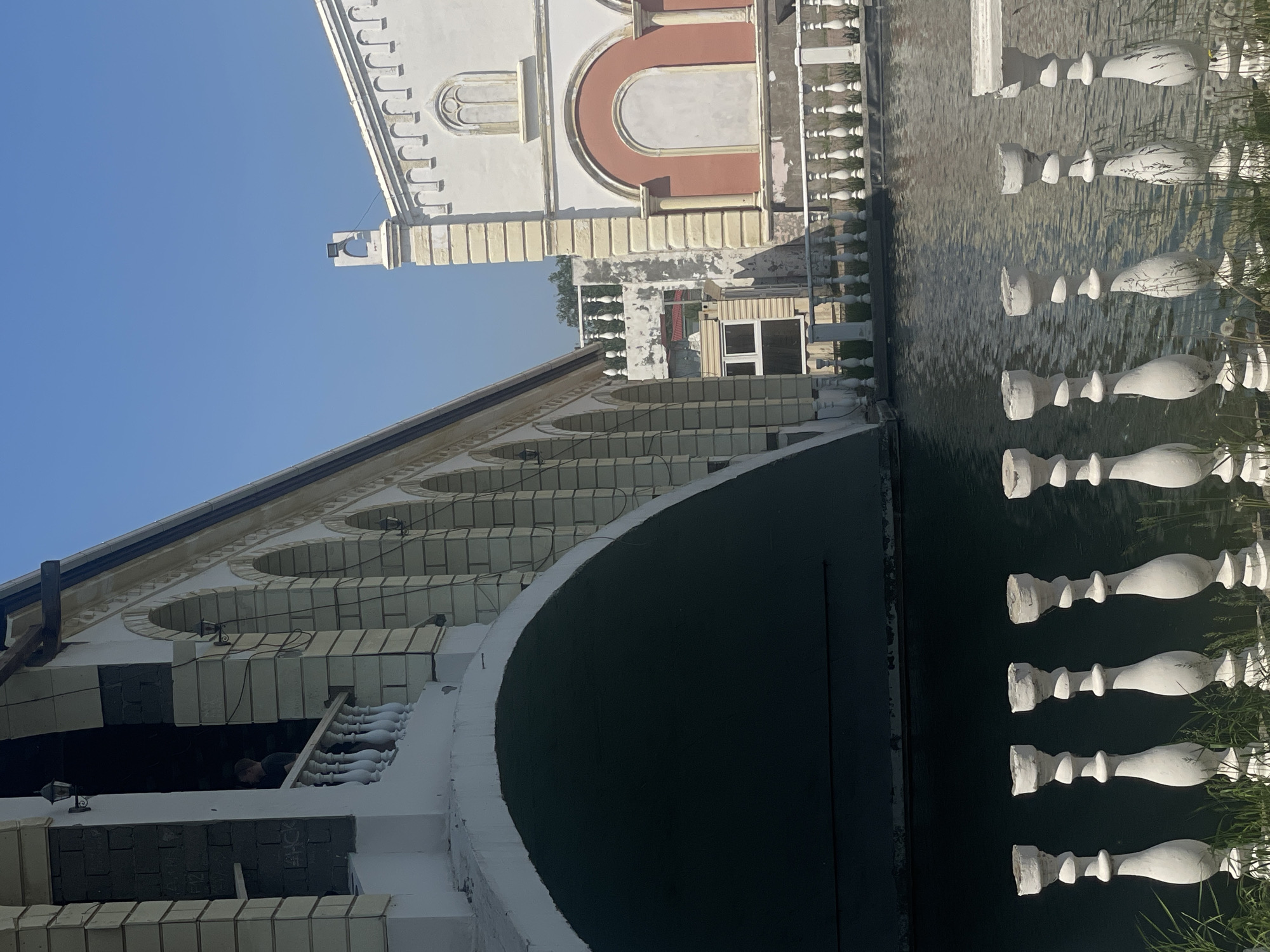 The image depicts an architectural complex featuring two ornate structures separated by a water channel, under a clear blue sky, in an unknown city in Russia.
On the left, a large, light-cream colored bridge or building spans the water with a prominent, dark archway. Its facade is characterized by a series of smaller arches supported by pilasters, with a white balustrade running along its upper edge. In an upper-level opening on the far left, a male figure in a dark t-shirt is visible, leaning on a white balustrade and looking towards the right. Exposed dark brickwork is visible around this opening. Several dark, wall-mounted lamps are affixed to the pilasters along the structure's side.
On the right, a white building with cream-colored pilasters features an arched and blank window on its upper section, and a larger, reddish-orange arched niche below it. Decorative molding lines the roofline, and a small architectural element, topped with an arch and a light fixture, extends from the roof.
A dark, rippled channel of water flows between the two structures, partially reflecting the sky. In the immediate foreground, a white balustrade composed of several ornate, weathered balusters with chipped tops stands, partially obscured by green grass and dandelions. The overall aesthetic suggests a classically inspired, possibly Venetian-themed, environment that shows signs of age or incomplete development.