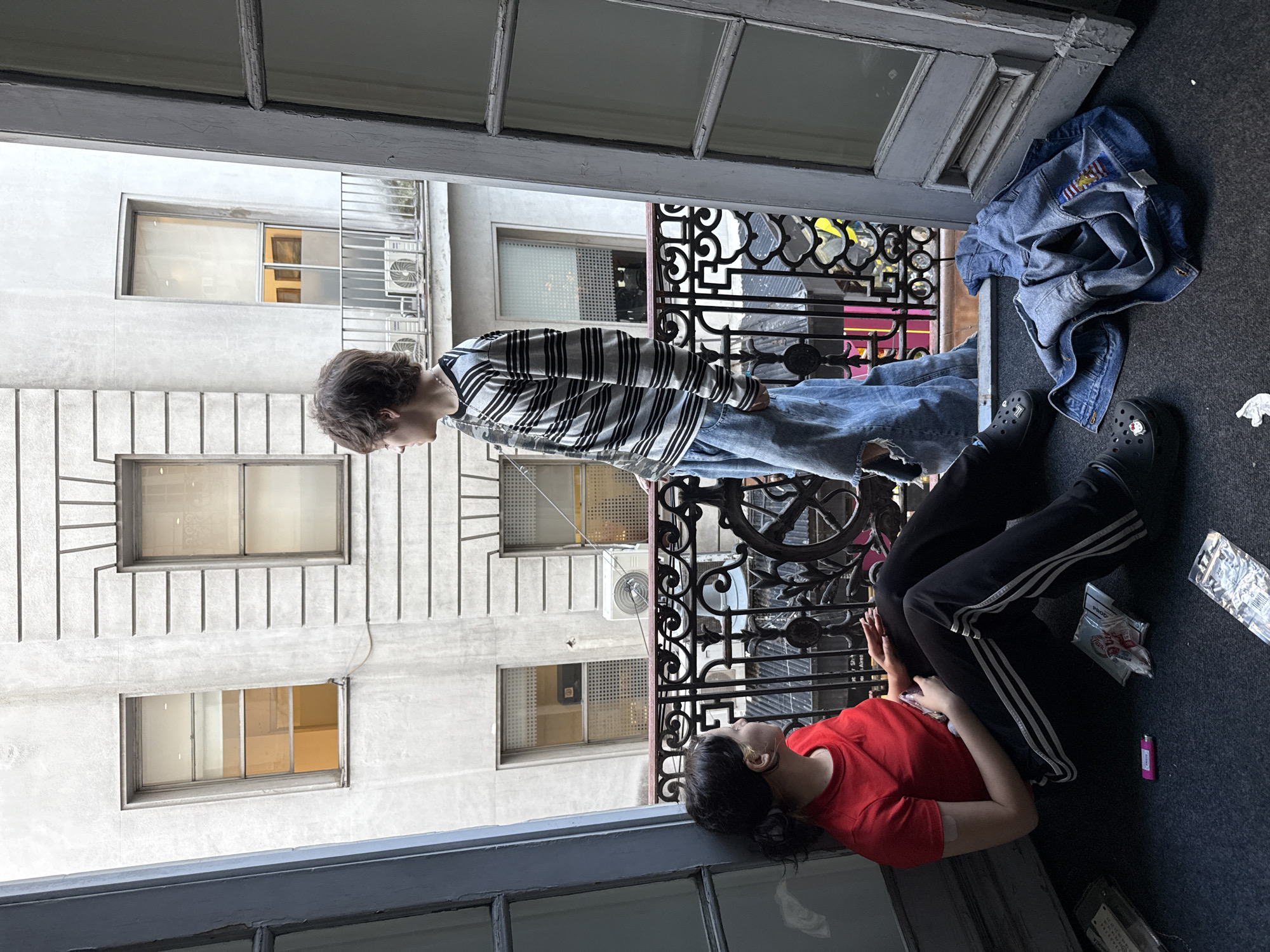 This image, set in an urban environment of Buenos Aires, Argentina, captures a view from an interior space looking out onto a small balcony and the facade of an opposite building.
Two individuals are present on the dark grey carpeted balcony. On the left, a person with short, dark, curly hair stands in profile, facing right and looking outwards. They are dressed in a black and white striped long-sleeved shirt, distressed light blue jeans, and black Crocs worn with white socks. A blue denim jacket, featuring a colorful patch that resembles a flag (with red, white, blue, and yellow elements), is lying on the balcony floor next to them.
On the right side of the balcony, a second person with dark hair tied back is resting on the floor, their head near the edge of the opening. This individual wears a plain red t-shirt and black track pants adorned with three white stripes down the sides, and has a small bandage visible on their left arm. Nearby on the floor are a purple lighter and a small clear plastic bag containing a white wrapper that appears to say "Flan" and "PROD."
The view across features a light-colored building with multiple windows, some fitted with metal security bars and several external air conditioning units. The balcony itself has an ornate black wrought-iron railing. The image appears to be taken during the daytime, under soft or overcast light conditions, suggesting a relaxed moment.