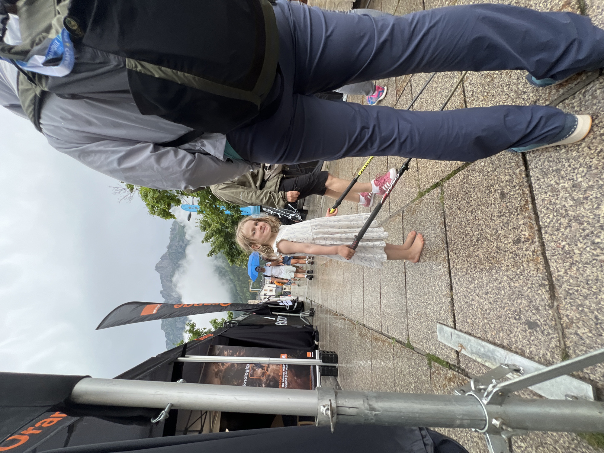 The image shows a cloudy day in Chamonix-Mont-Blanc, France, with a group of people gathered on a wet paved walkway. In the foreground, a young girl in a white dress is barefoot and holding ski poles. To her right, the lower half of a person wearing dark blue pants and a light gray jacket is visible. Behind the girl, several other people are walking along the path, one of whom is holding a blue umbrella. Tents and banners, one with the "Orange" logo, are set up along the side of the walkway. In the background, mist partially obscures the view of the mountains.