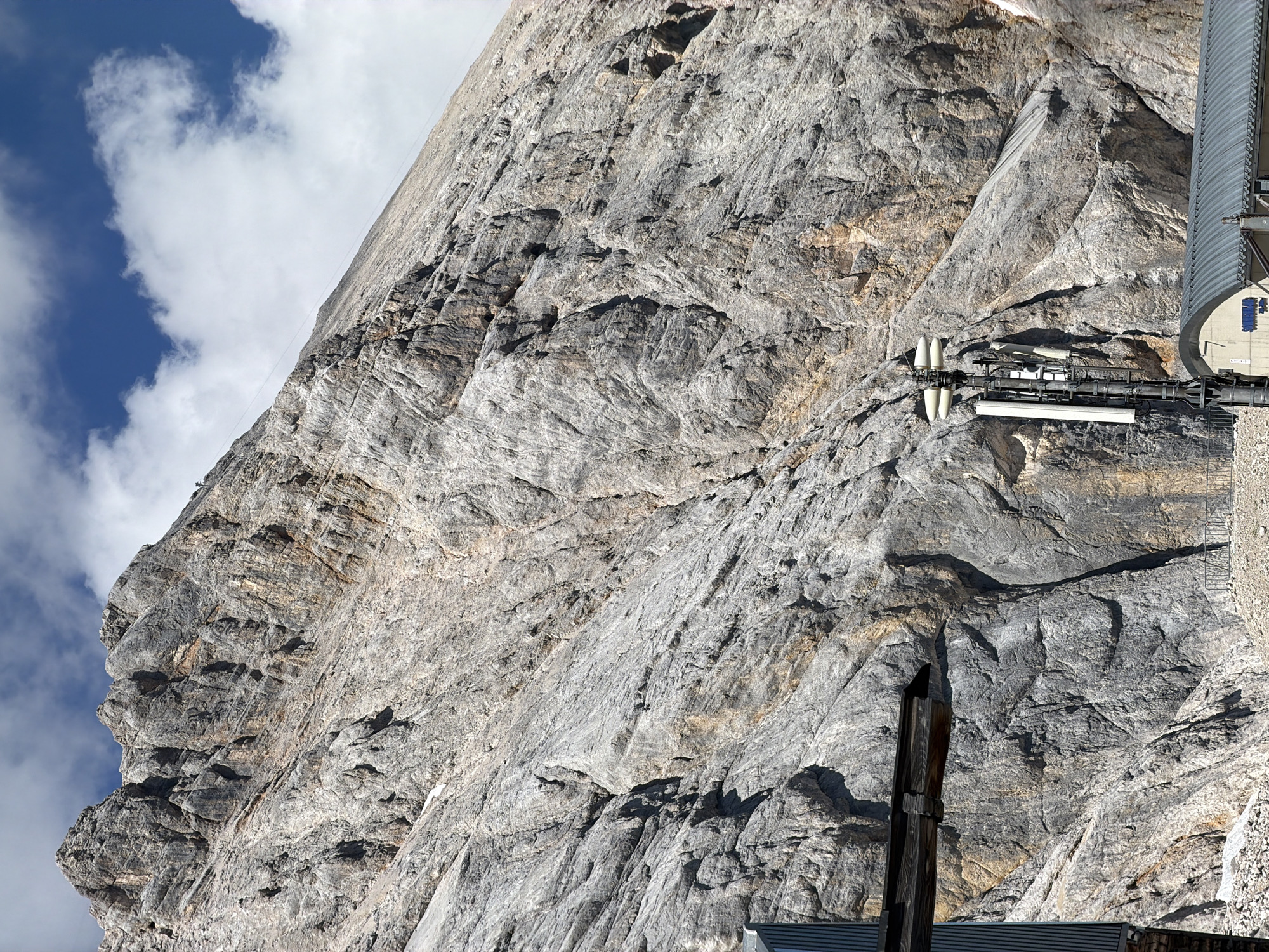 This image captures a dramatic, wide-angle view of the Zugspitze mountain in Garmisch-Partenkirchen, Germany. The mountain face dominates the frame, its rugged, gray rock surface textured with fissures and geological formations. Sunlight highlights the rough terrain, casting shadows that accentuate the mountain's imposing scale. The sky above is a brilliant blue, dotted with fluffy white clouds. In the foreground, a metallic structure, possibly part of a cable car station or observation post, is visible on the left, featuring wooden beams and corrugated metal. To the right, a tall communication tower with multiple antennas stands prominently against the mountain backdrop, situated in front of a building with a curved, metal roof. The lower portion of the image shows a rocky, uneven ground, suggesting a high-altitude, natural environment.