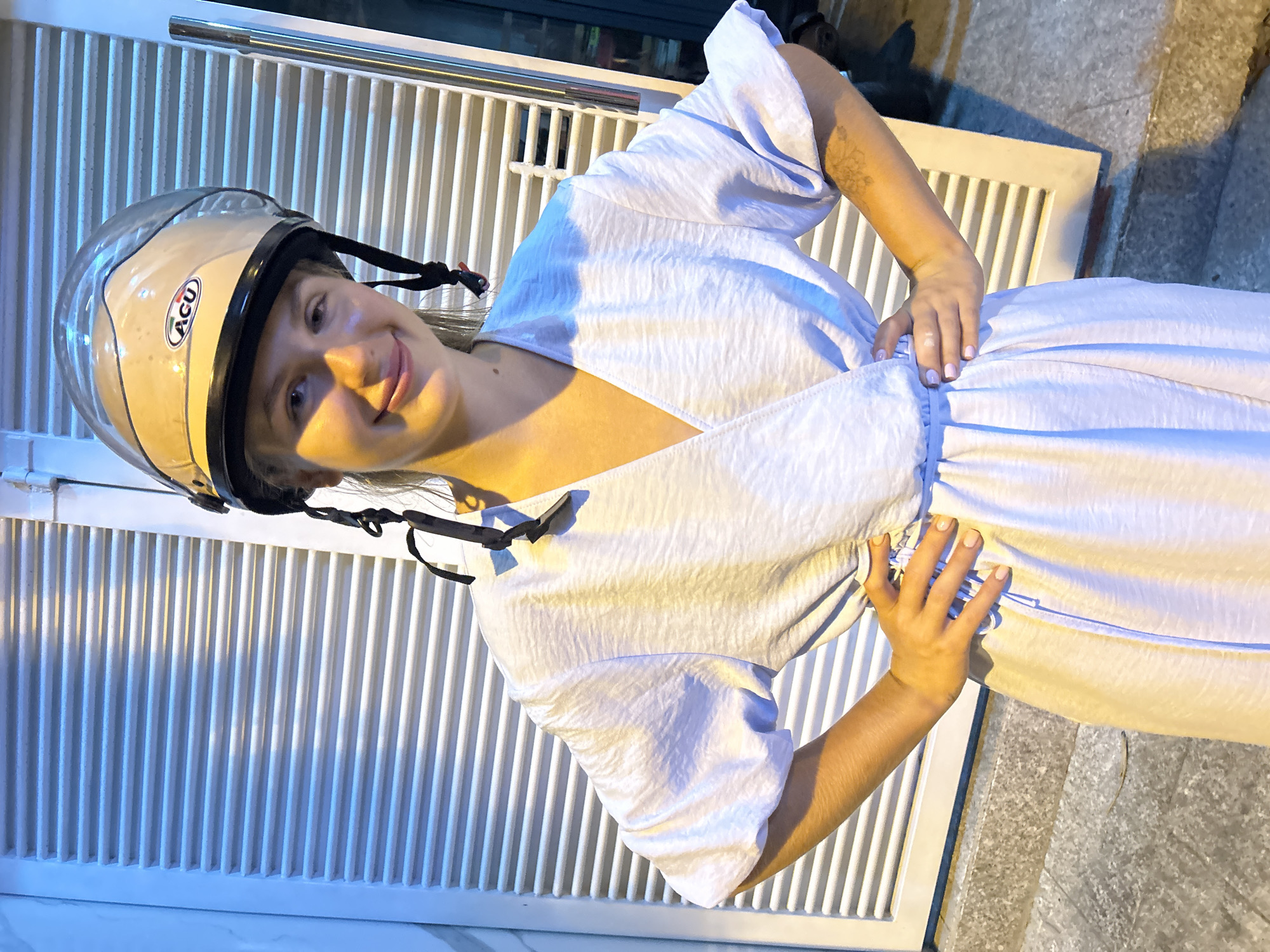 A woman wearing a beige and clear helmet with a black chin strap stands in front of a white, slatted gate. She is wearing a light blue, textured dress with short, puffed sleeves and a V-neck. Her arms are bent, and her hands are placed on her hips, one hand near the hem of her dress. She has light skin and is smiling with her tongue slightly visible. There is a tattoo on her upper arm, and her fingernails are painted a light pinkish-lavender. The lighting suggests it is evening, possibly with artificial streetlights illuminating the scene. The background consists of the gate, a textured wall, and a concrete floor with a dry leaf. The overall mood is casual and friendly. The helmet has a sticker that says "ACU".