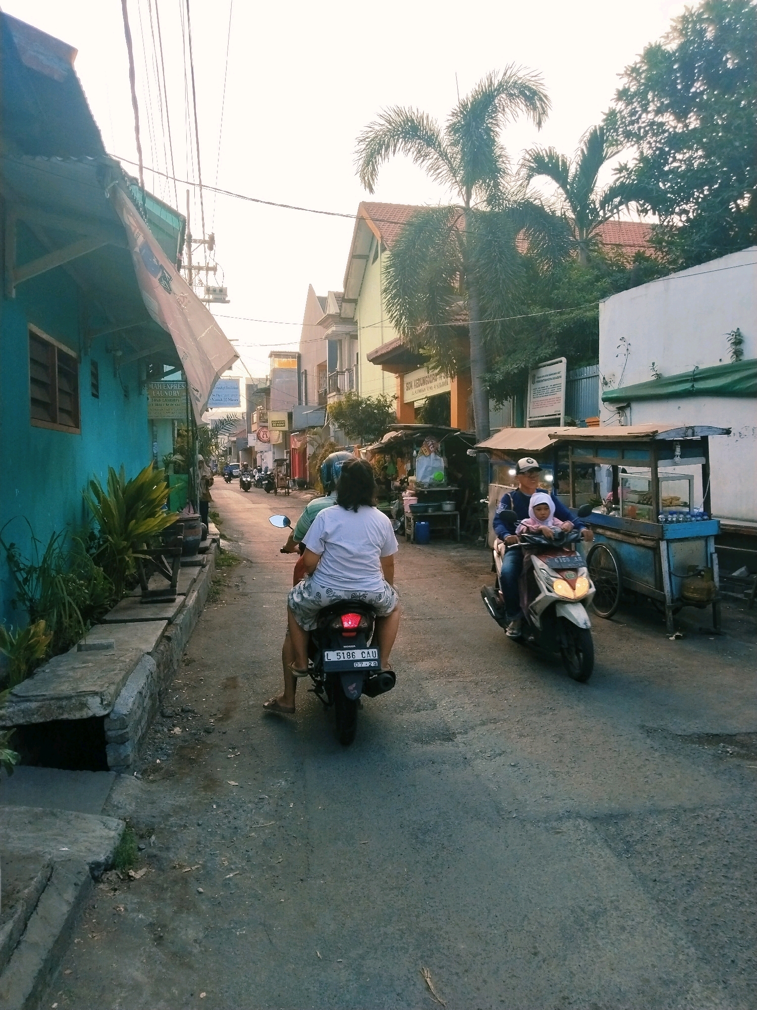 In a narrow street in Surabaya, Indonesia, two motorbikes are moving in the same direction. On the left, a person is riding a motorbike, with another person riding pillion. The rider is wearing a white t-shirt and patterned shorts, and the pillion rider has dark hair and is wearing a light-colored t-shirt. The motorbike has a license plate that reads "L 5186 CAU". On the right, a man is riding a motorbike with a child seated in front of him. The man is wearing a dark blue long-sleeved shirt, dark pants, and a cap. The child is wearing a pink headscarf and light-colored clothing. The motorbike has a license plate that reads "L 6151 AA".
The street is lined with buildings on both sides. On the left, a turquoise-colored building with windows and an awning is visible, with lush green plants growing along its base. On the right, there are several stalls or small shops, one with blue-trimmed structures and what appear to be food vendors. In the background, more buildings and palm trees can be seen, suggesting an urban or semi-urban setting. Overhead, electrical wires crisscross the sky. The overall lighting suggests it is either early morning or late afternoon.