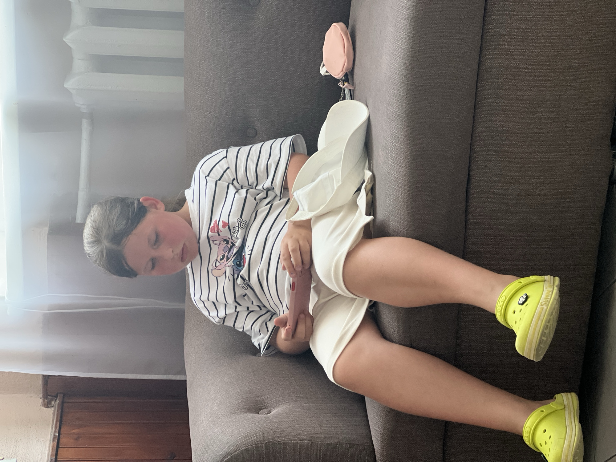 A young girl, with her hair pulled back in a ponytail, sits on a gray sofa in what appears to be an indoor waiting area or room. She is wearing a horizontally striped t-shirt with a cartoon character print and light-colored shorts. Her feet are adorned with bright yellow clogs, and she is holding a pink mobile device in her hands, looking down at it. To her right, on the sofa, rests a white hat and a small pink pouch. The background shows a wooden wall paneling on the left, a radiator and window with sheer curtains on the right, suggesting an interior setting. The overall scene is calm and ordinary, depicting a child engaged with a personal device. The location is specified as Сонячне, Ukraine.