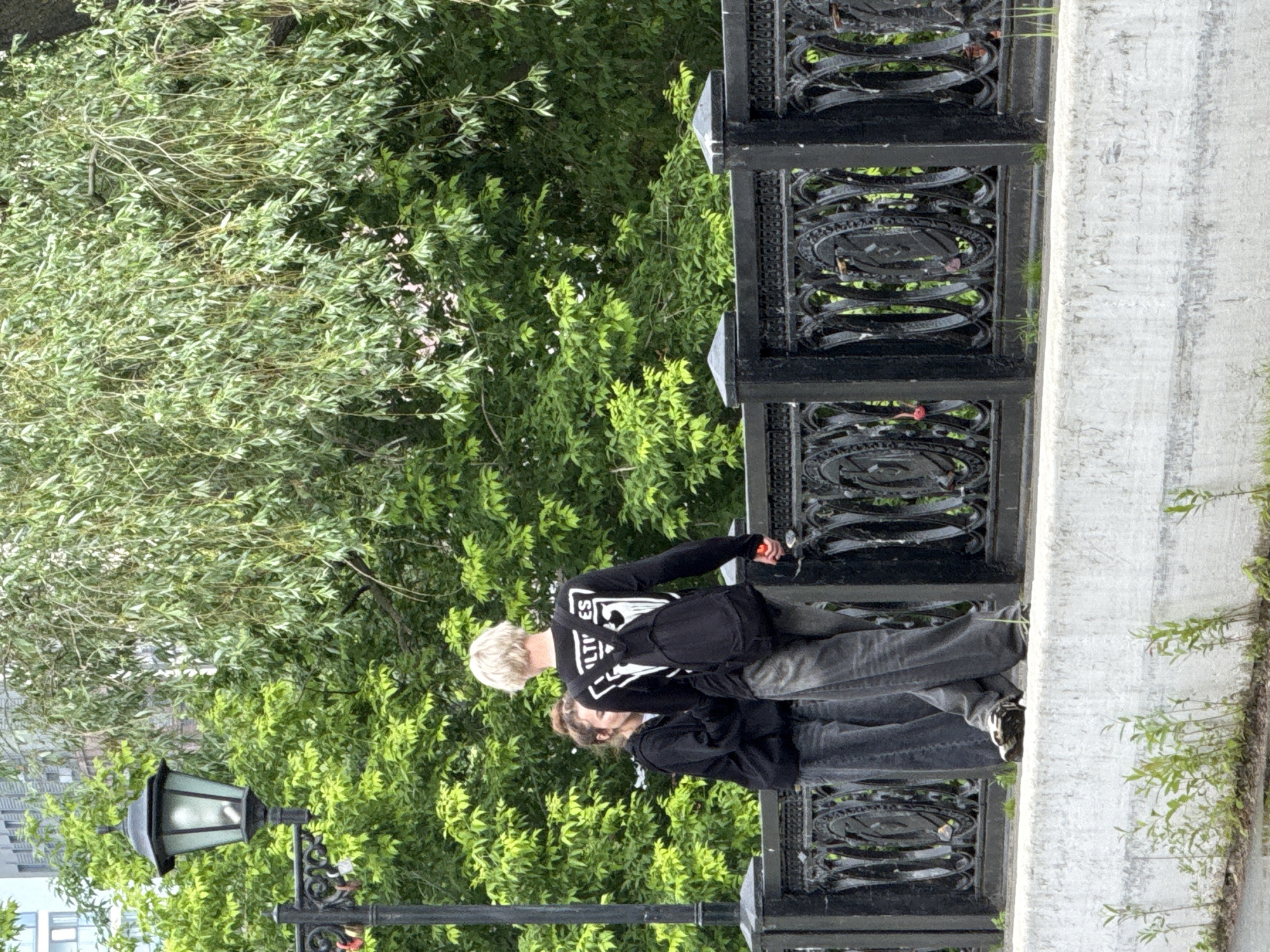 Two individuals, one with blonde hair and the other with brown hair, are standing on a bridge. The blonde-haired person, wearing a black long-sleeved shirt with white lettering and a backpack, is facing away from the camera and holding a red object in their right hand. The brown-haired person, wearing a black hoodie, is facing left, partially obscured by the blonde-haired person. They are both dressed in casual clothing, including denim jeans.
The bridge has a black decorative metal railing with circular motifs. Behind the railing, lush green foliage from trees is visible. To the left of the individuals, a black lamppost with a lantern stands. The bridge is constructed with a light-colored, textured concrete barrier at the bottom. Small green plants are growing along the base of the concrete barrier. The scene appears to be an outdoor urban setting, likely a park or pedestrian area within the city of Izhevsk, Russia. The overall lighting suggests an overcast or diffused daylight condition.