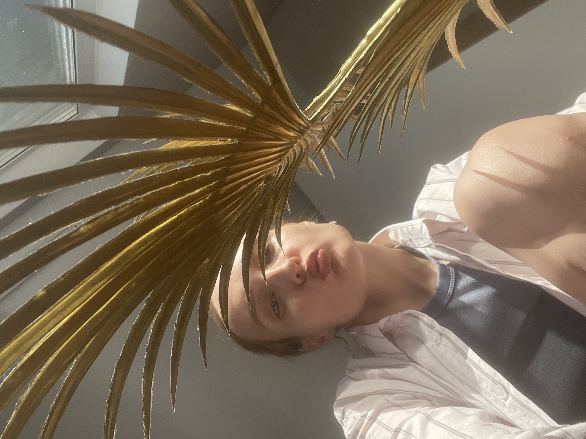 A low-angle shot presents a person, possibly female, with their face partially obscured by a gold-colored palm frond. The person has their lips pursed. They are wearing a blue tank top and a white striped shirt. The background shows part of a window and a gray wall. The location in Sepych, Russia is unverifiable from the image.
