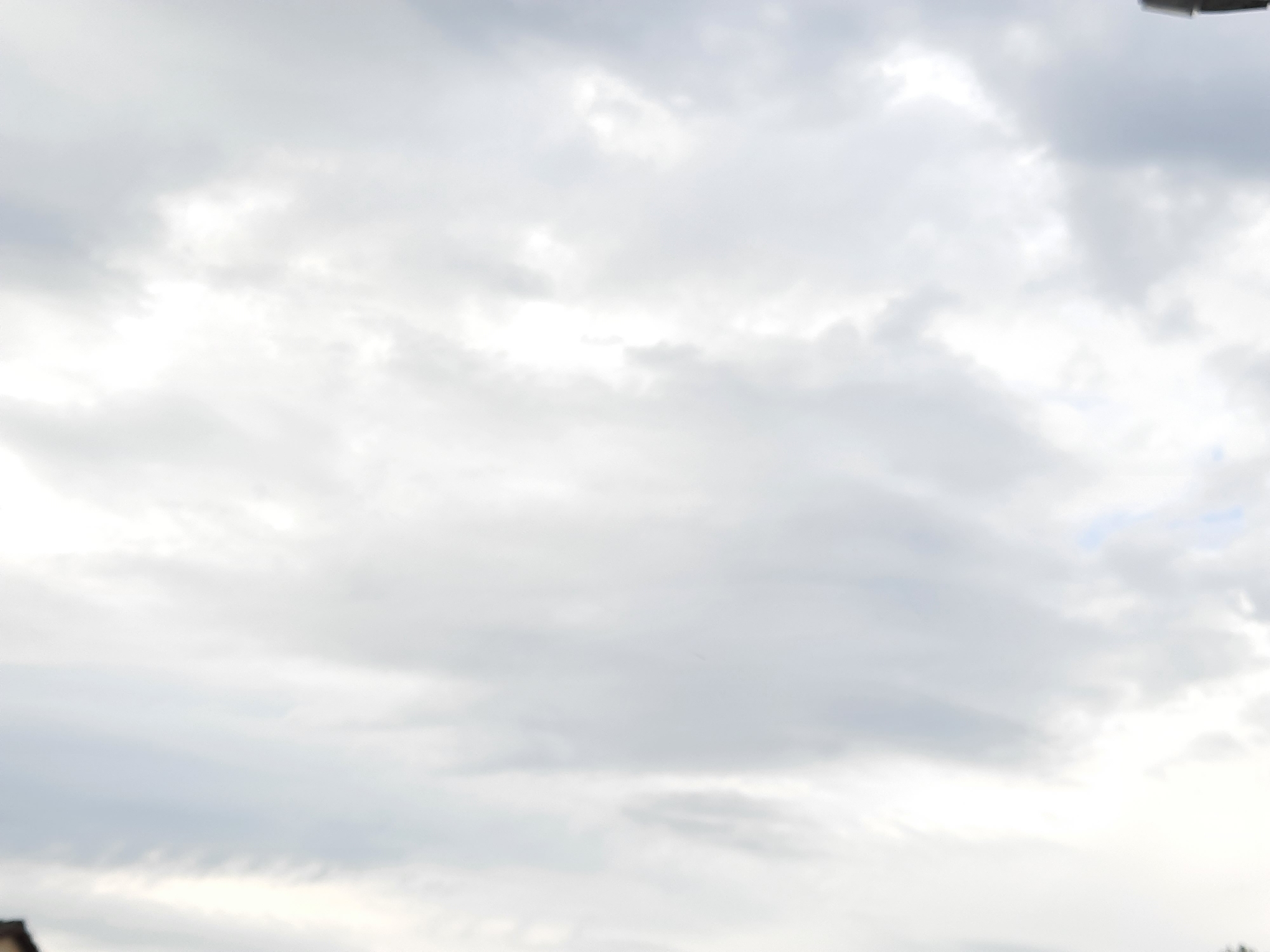 The image shows a cloudy sky with some bright areas indicating the sun trying to break through. The bottom left corner of the image shows a dark object which appears to be the edge of a building or structure. There are no discernible people, subjects, or actions captured in this image. The scene is abstract and solely focused on the atmospheric conditions and a hint of terrestrial structure.