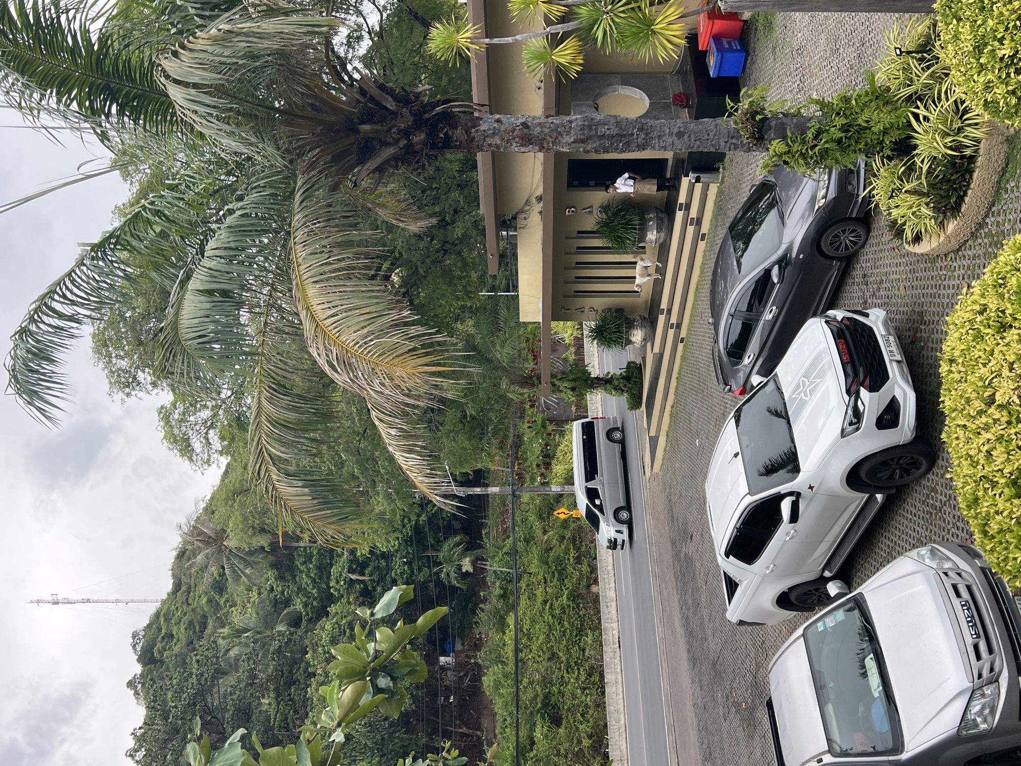 The image shows a street scene in Baan Chaweng Noi, Thailand. A building with multiple steps is present; a person is standing in front of it. A gray car and two white pickup trucks are parked on the paved area in front of the building. A palm tree overhangs the building and parking area. Across the road, a hill covered in vegetation is visible. A white van is driving on the road. A communications tower is visible in the distance. The sky is overcast.