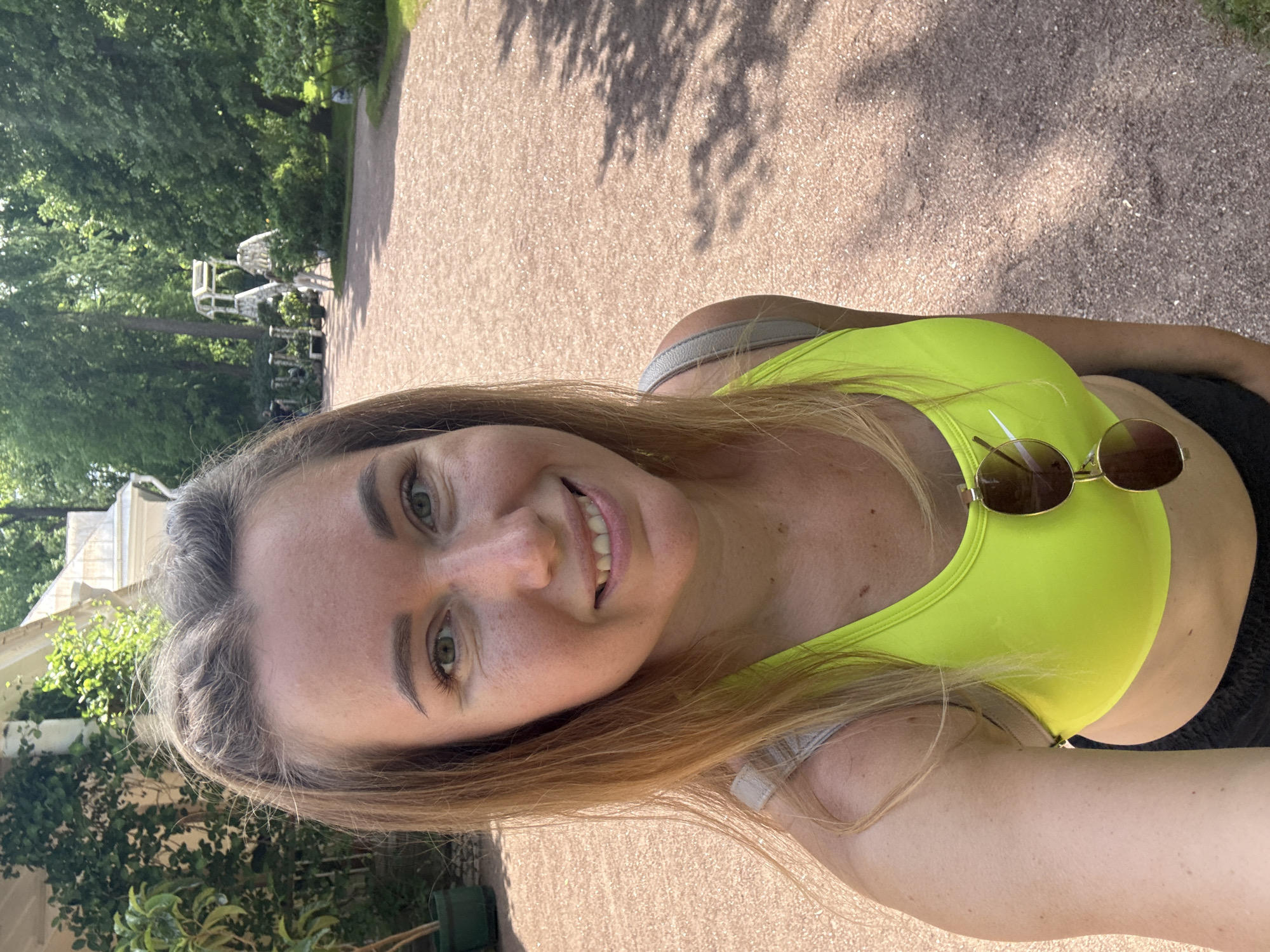 A fair-skinned woman with light brown hair, pulled back from her face, smiles directly into the camera in a selfie taken outdoors on a sunny day. She has green eyes and visible freckles on her face and shoulders. She is wearing a neon yellow sports bra, from which brown-tinted sunglasses hang. A light-colored strap, possibly from a bag, is visible over her left shoulder. She is also wearing dark bottoms, likely shorts or leggings. The background features a wide, reddish-brown gravel or compacted earth path with shadows cast across it. Lush green trees and foliage flank the path. In the mid-background, several white, decorative garden structures, possibly arches or trellises, are visible amongst the trees. A light-colored building with a roof is partially visible behind greenery on the far left. The specific location within the city of Unknown, Unknown is not identifiable from the image.