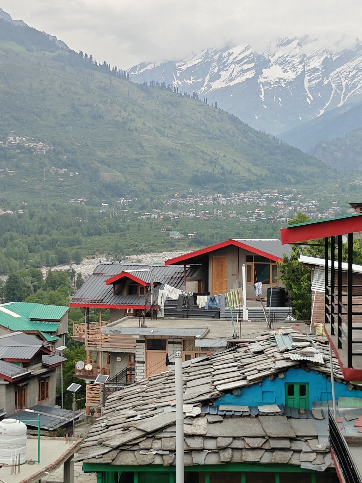 The image depicts a tiered settlement in the city of Vashisht, India, against a backdrop of mountainous terrain under an overcast sky. In the far background, majestic snow-capped mountains are partially obscured by clouds. Below these, a vast expanse of green, forested mountains slopes downwards, dotted with numerous smaller settlements. A wide, rocky riverbed with a visible water flow meanders through the valley at the base of these mountains.
In the mid-to-foreground, a dense cluster of multi-story buildings forms the settlement. Rooftops exhibit a variety of materials and styles:
* Several buildings feature dark gray corrugated metal roofs with distinct red eaves and gable ends. On one such roof, laundry is visible hanging on clotheslines.
* Other structures have traditional roofs constructed from overlapping, irregular gray slate or stone tiles. One building in the lower right foreground displays a bright blue gable end with a small green double-door window, partially covered by these slate tiles over a corrugated metal base.
* Another building has a bright green corrugated metal roof.
The buildings appear to be constructed with concrete walls and incorporate wooden elements for doors, windows, and balconies. Specific objects visible on and around the rooftops include:
* White plastic water storage tanks.
* Clotheslines with various items of laundry hanging to dry.
* Two pole-mounted solar panels, possibly for street lighting, along with two satellite dishes positioned nearby.
* Wooden railings and support structures for balconies.
* Two dark birds, possibly pigeons, are perched on a dark gray roof in the middle ground.
No human figures are visible within the frame. The overall scene conveys a blend of traditional mountain architecture and modern amenities amidst a rugged natural landscape.