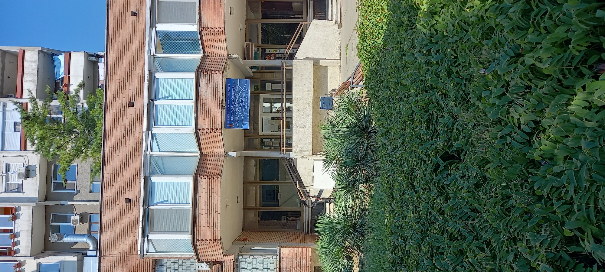 The image shows the facade of a multi-story building with red brick accents, located in the city of Silistra, Bulgaria. The building features several floors with windows, some of which have blinds. A blue banner with text in Bulgarian hangs above the entrance. The ground level has glass doors and windows, with a ramp leading up to the entrance. In front of the building, there is a dense area of green foliage, including yucca plants and a lower shrub layer. In the background, a taller apartment building is visible, with a tree growing in front of it. The sky is clear and blue.