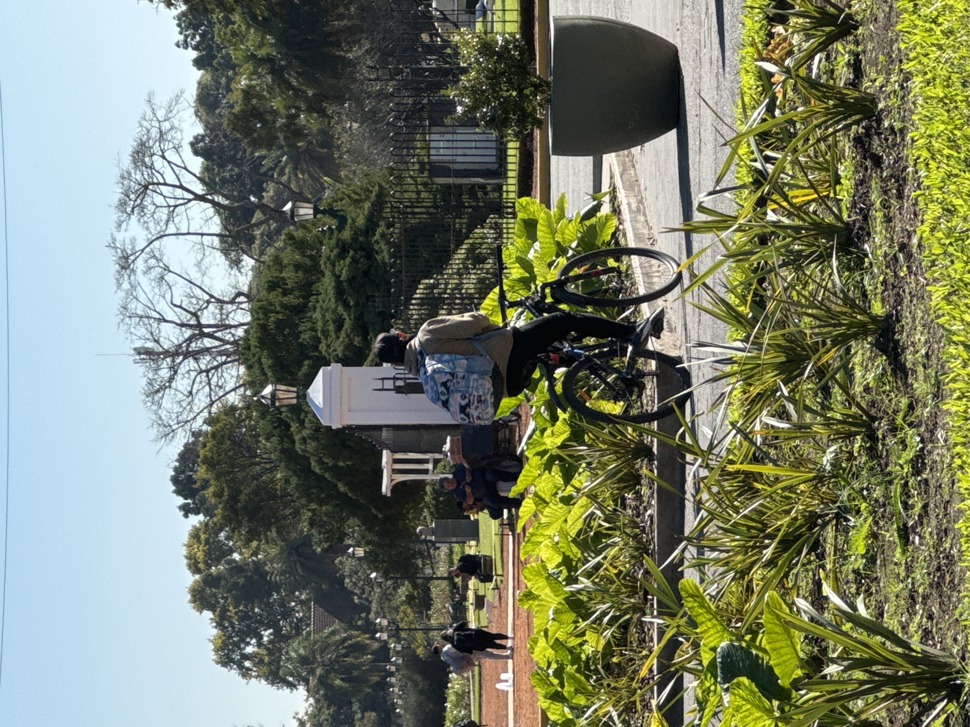 The image depicts a scene in Buenos Aires, Argentina. A person with a backpack is sitting on a bicycle on a paved path. The path is lined with plants and greenery. In the background, there's a white structure, possibly a monument or building, along with trees and a fence. Other people are walking in the distance. The weather appears to be sunny and clear.