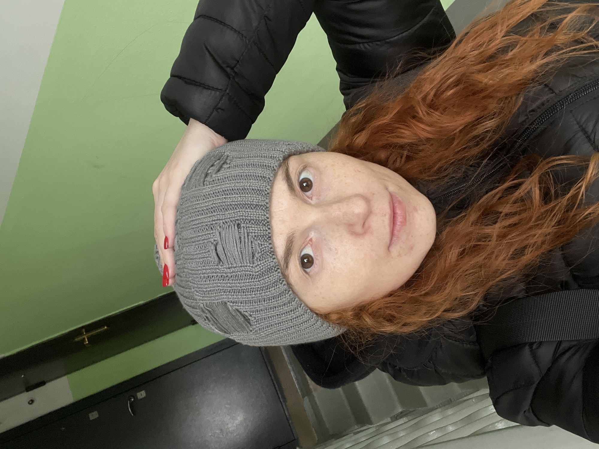 A close-up, vertically oriented indoor portrait captures a young woman in Chaikovsky, Russia, wearing cool-weather attire. The woman has long, wavy red hair and a fair complexion with numerous freckles across her face. She wears a grey knitted beanie hat, which features decorative rips or slits, and a black puffer jacket. Her brown eyes are wide, and she looks directly at the camera with a neutral to slightly surprised expression. Her right hand, adorned with red nail polish, is visible at the top of the frame, resting against her beanie. The background consists primarily of a plain light green wall, which meets a darker, possibly grey or black, horizontal surface or ledge towards the bottom of the frame. An indistinct dark object is visible in the lower left corner, possibly part of a door or furniture. No text is visible in the image.