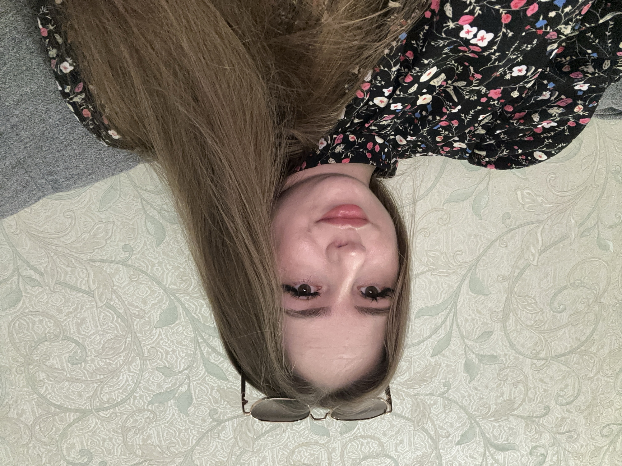 A young woman with long brown hair and glasses on top of her head is pictured from the chest up, looking at the camera. She is wearing a black dress with a floral pattern. The background is a wallpaper with a light floral design. The scene appears to be indoors, possibly in a home.