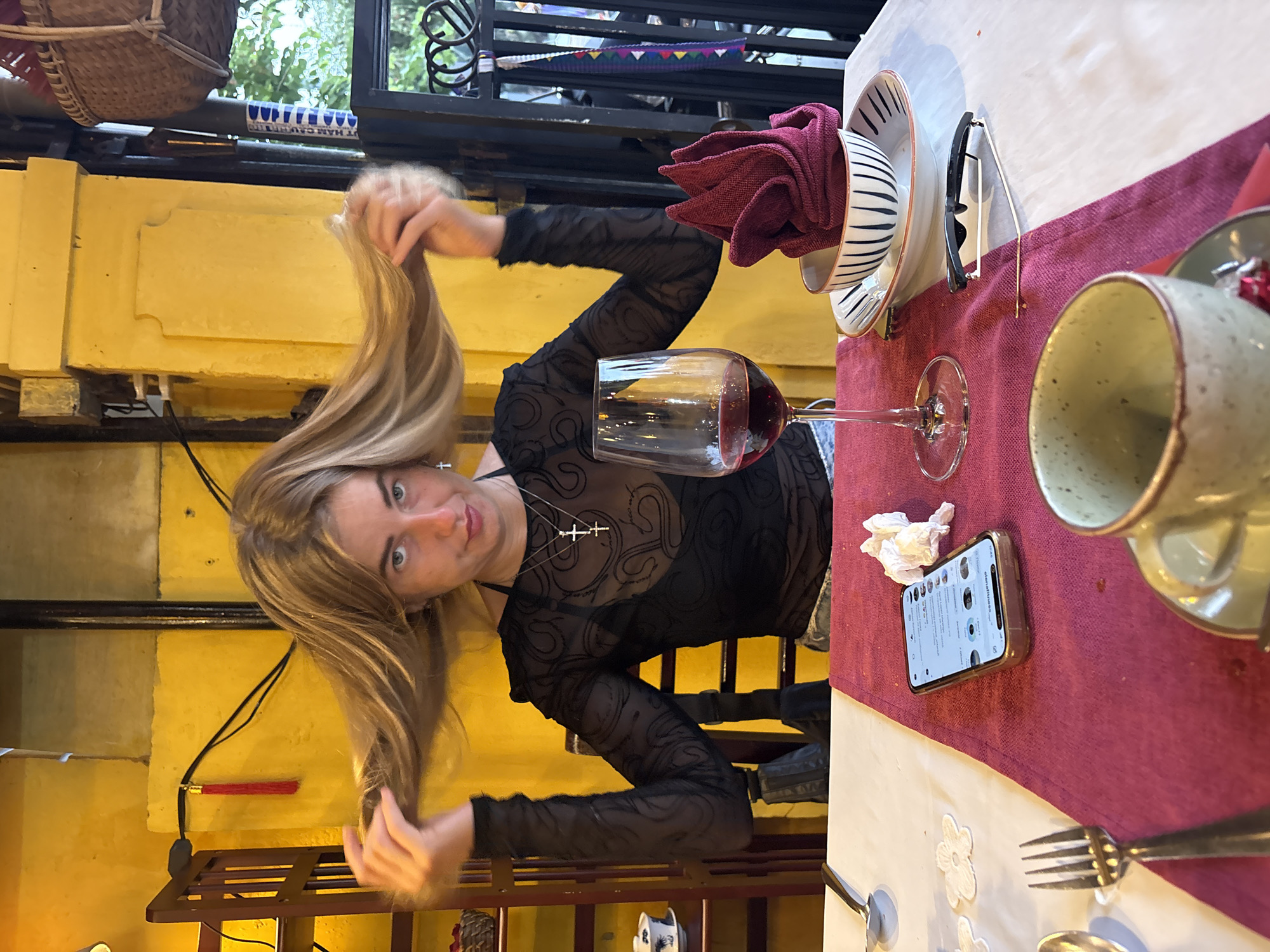 The photograph captures a blonde-haired woman in a black sheer top, holding her hair in her hands. She is seated at a table set for dining, marked by a red tablecloth, plates, a glass of red wine, a napkin, and silverware. A smartphone rests on the table. The environment appears to be a restaurant or dining space in Hội An Ward, Vietnam, characterized by its yellow wall and architectural features visible in the background.