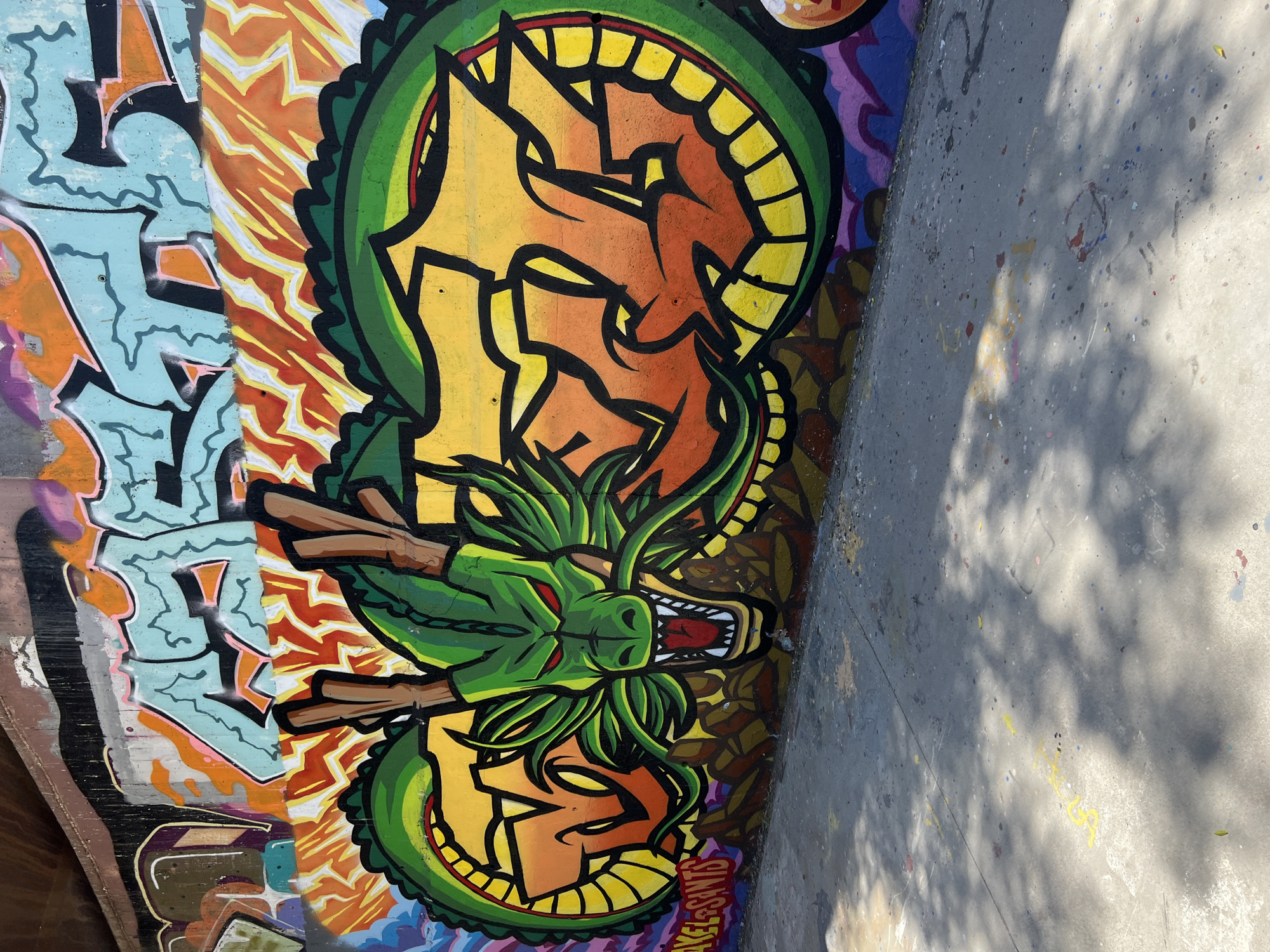 This is a photograph of a large graffiti mural on a concrete wall, likely in an urban outdoor setting such as a skatepark or underpass, given the presence of the concrete ground with paint splatters and shadows from overhead foliage. The central subject of the mural is a stylized green dragon head with red eyes and an open mouth, revealing sharp teeth. The dragon is depicted with a mane of green tendrils and two brown horns. To the right of the dragon's head, within its coiled body, are bold, abstract graffiti letters filled with a gradient of yellow and orange. The background of the mural features a vibrant array of colors, including orange and yellow jagged patterns resembling flames, and sections of turquoise and purple lettering. The lower left corner of the mural shows the signature "AKEL.SAINTS" in red and white spray paint. The ground in front of the wall is concrete, with numerous small splatters of paint in various colors, and dappled sunlight creating shadows across the surface. The concrete wall itself appears weathered. The overall impression is one of a dynamic and colorful street art piece.