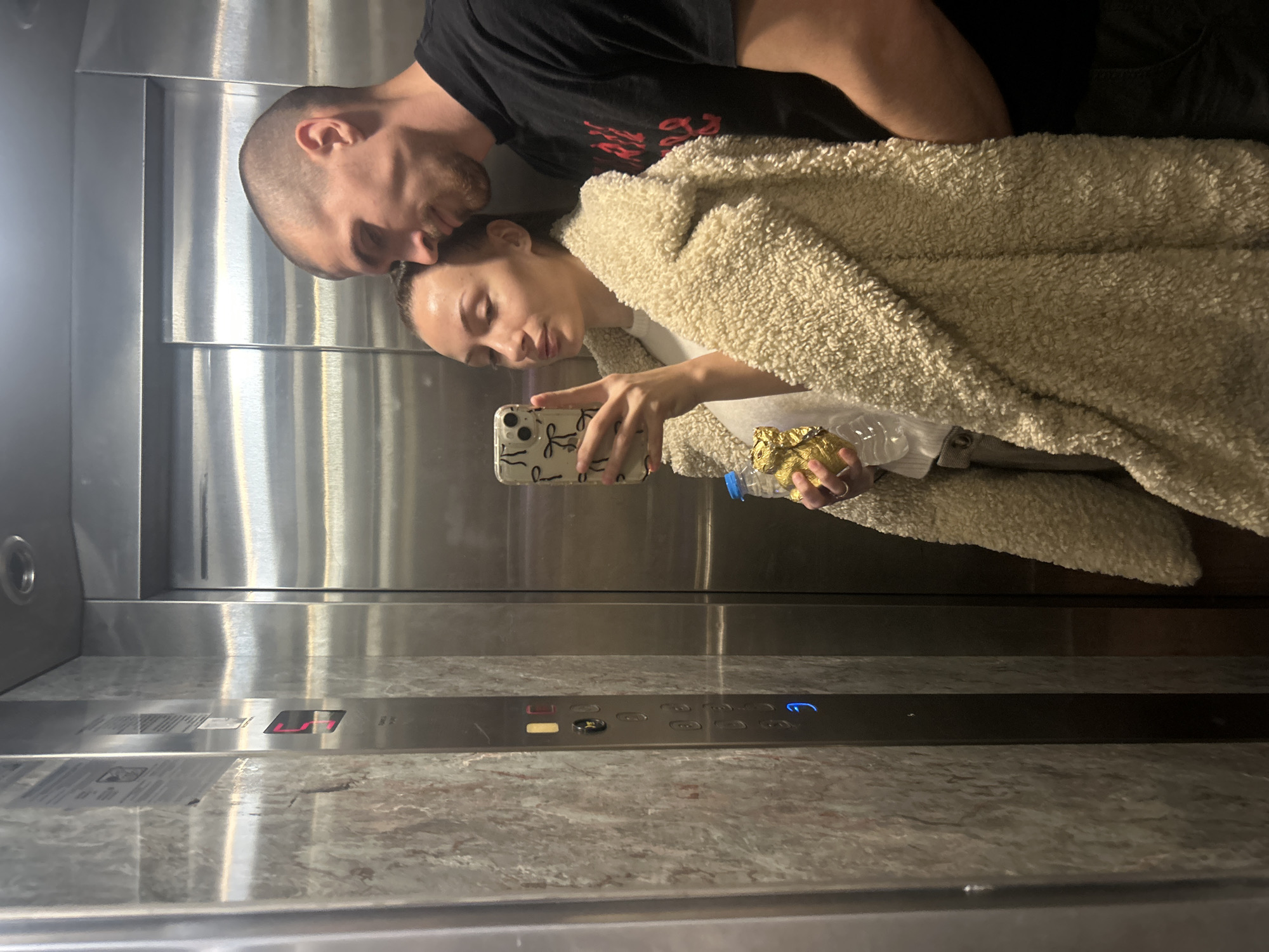 A man and woman are taking a selfie in an elevator. The man, with a shaved head and beard, is wearing a black t-shirt and stands behind the woman, his head leaning toward hers. The woman is wearing a light-colored, textured coat and holding a mobile phone in her right hand, with a water bottle and what appears to be a chocolate wrapper in her left. The elevator has a stainless steel wall, and a control panel with buttons is visible below the couple. The floor of the elevator is gray with a speckled pattern. The location is Burgas, Bulgaria, based on the elevator design which is found in hotels and businesses there.