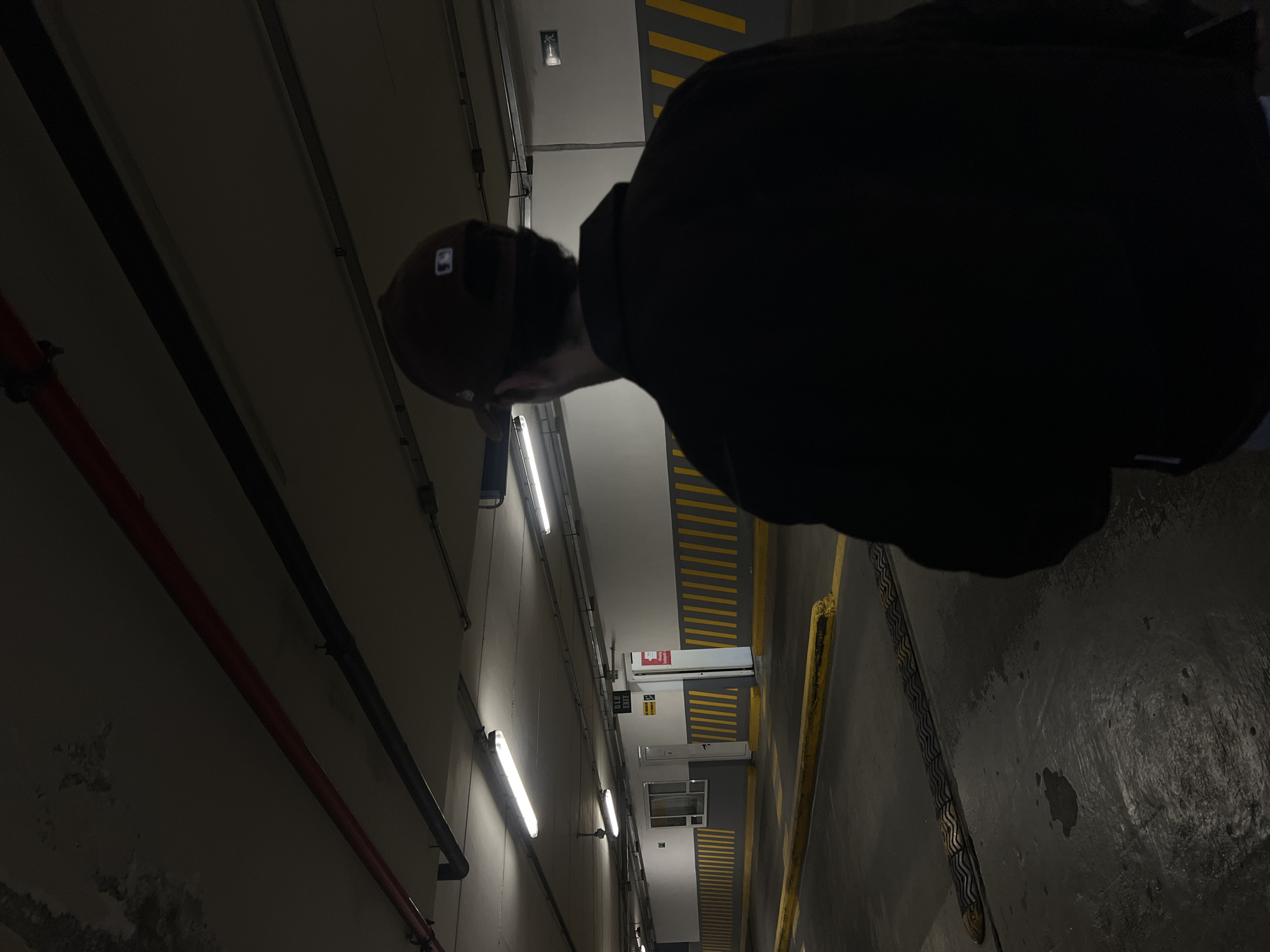The image shows a person wearing a baseball cap and a dark jacket in what appears to be an underground parking garage in Yerevan, Armenia. The walls are grey and illuminated by fluorescent lights. There are red pipes running along the left wall. Yellow lines are painted on the floor and walls. A chain lies on the ground. A sign is visible in the background.