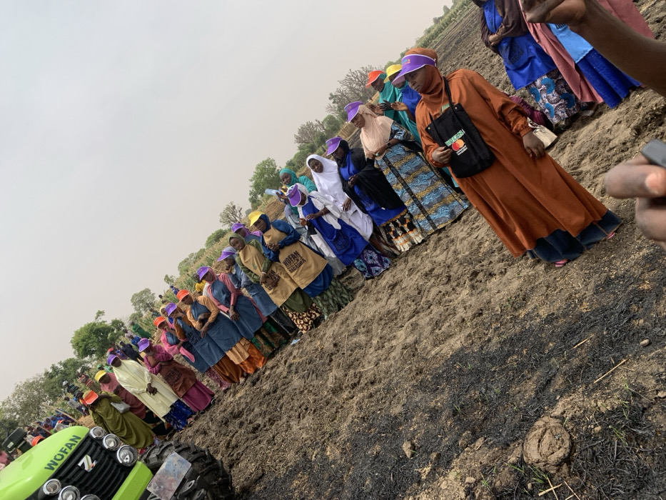A group of women are gathered on a field in Ringimawa, Nigeria. They are standing in a line, facing the camera. They are wearing colorful clothing. Some of them are holding yellow bags with the words "WOMAN IN AGRICULTURE" printed on them. There is a green tractor in the background. The field is brown and dry, and there are some charred patches of vegetation. The women are listening attentively as someone speaks. The sky is overcast. The coordinates are: Latitude: 11.666, Longitude: 8.414. This appears to be an agricultural training event or a demonstration. The presence of the tractor and the phrase "WOMAN IN AGRICULTURE" on the bags suggests an initiative focused on empowering women in agriculture.