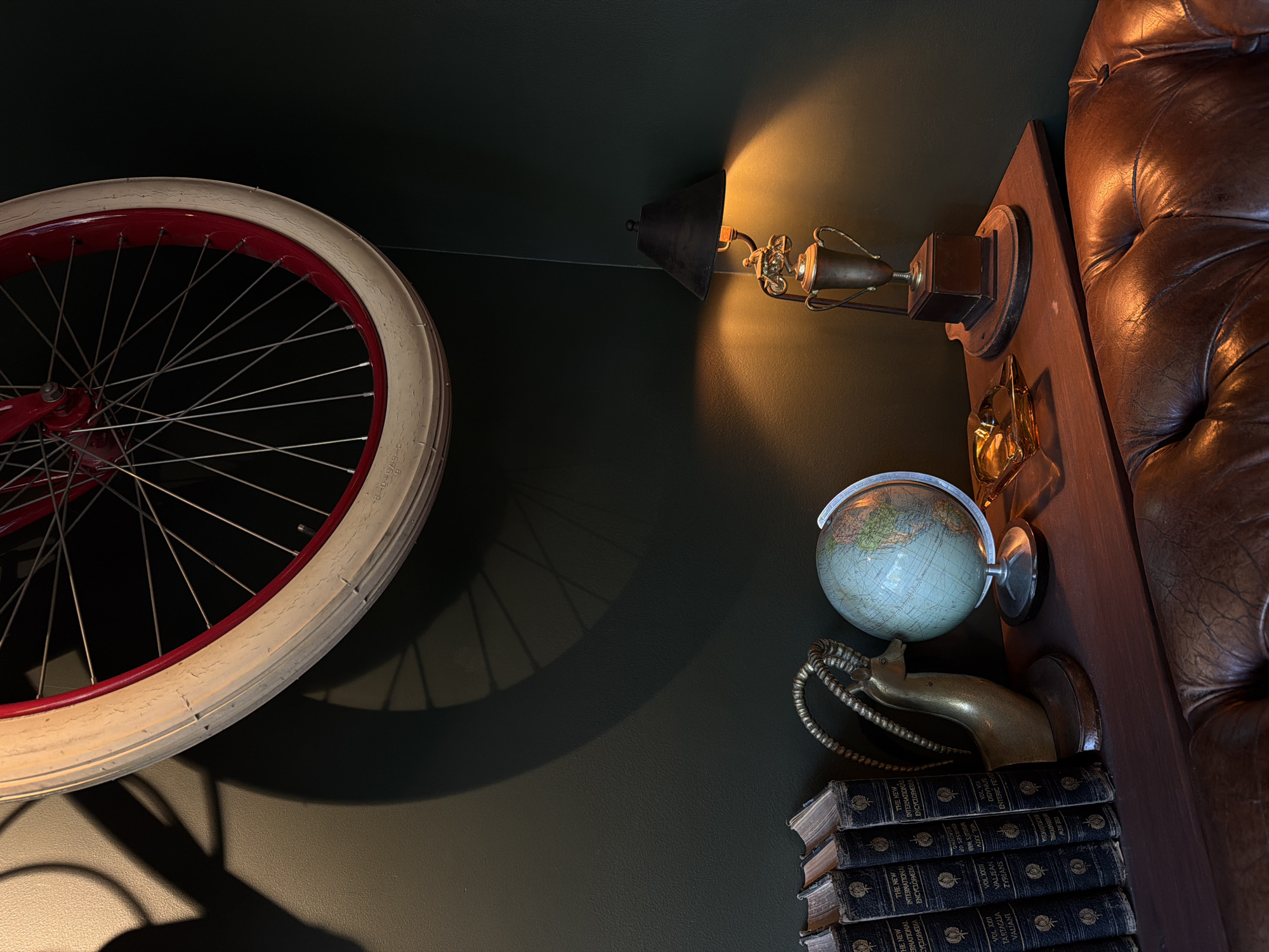 The image shows a collection of objects arranged on a dark green wall next to a tufted brown leather sofa. A red bicycle wheel with a white tire is mounted on the wall to the left. Next to it, there is a lamp illuminating a small gold trophy mounted on a wooden block. Below the lamp, there is a small globe positioned above a golden goat head with a linked chain around its neck. A stack of old encyclopedias is placed under the goat head. The location of the scene is West Hollywood, United States of America.