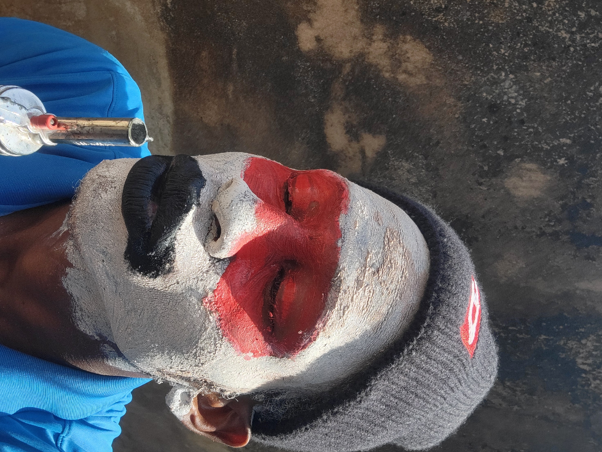 An individual with face paint is being sprayed with an unknown substance from a metal canister. The face paint is white with black markings around the forehead and red around the eyes. The person is wearing a gray knit cap with a small red and white symbol on the side and a blue shirt. The background is a mottled gray surface. Based on the location and the cultural context, it is highly probable this scene is taking place during the masking season festival in Nkpor, Nigeria.