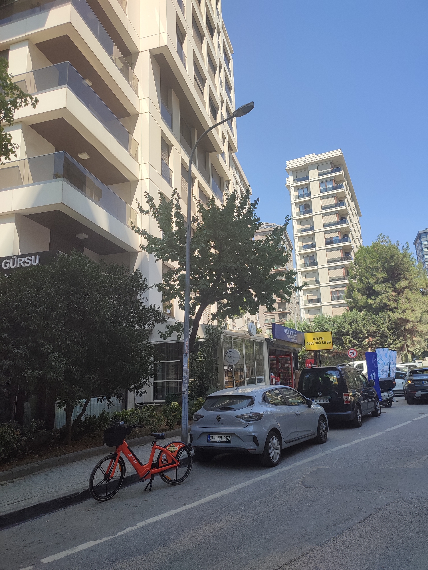 The image depicts a street scene in Kadıköy, Turkey. In the foreground, an orange electric bicycle with "tripy" branding is parked on the sidewalk. Behind it, a light grey hatchback car is parked on the street, with a license plate that reads "34 PMM 362". Further down the street, a black van and other vehicles are parked. The street is lined with modern buildings, including a tall apartment building on the left and a narrower, taller building in the background on the right. Trees and foliage are visible along the sidewalk and in front of the buildings. The sky is clear and blue. A street lamp is also visible. There are signs on some of the buildings and in front of a small storefront.