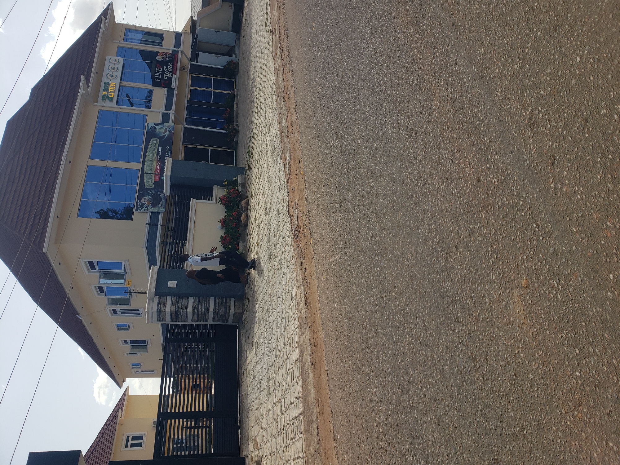 This is a medium-angle, eye-level, outdoor, full shot of a two-story building on a sunny day, likely in the city of Gura Top, Nigeria. The building is painted a pale yellow and features a brown, shingled roof. Large, reflective blue windows dominate the second floor. A black metal gate is visible on the left, leading to a paved walkway. Two individuals are walking on the walkway in front of the building. The foreground is occupied by a paved road. Signs are visible on the building, including one that says "FINE Wine."