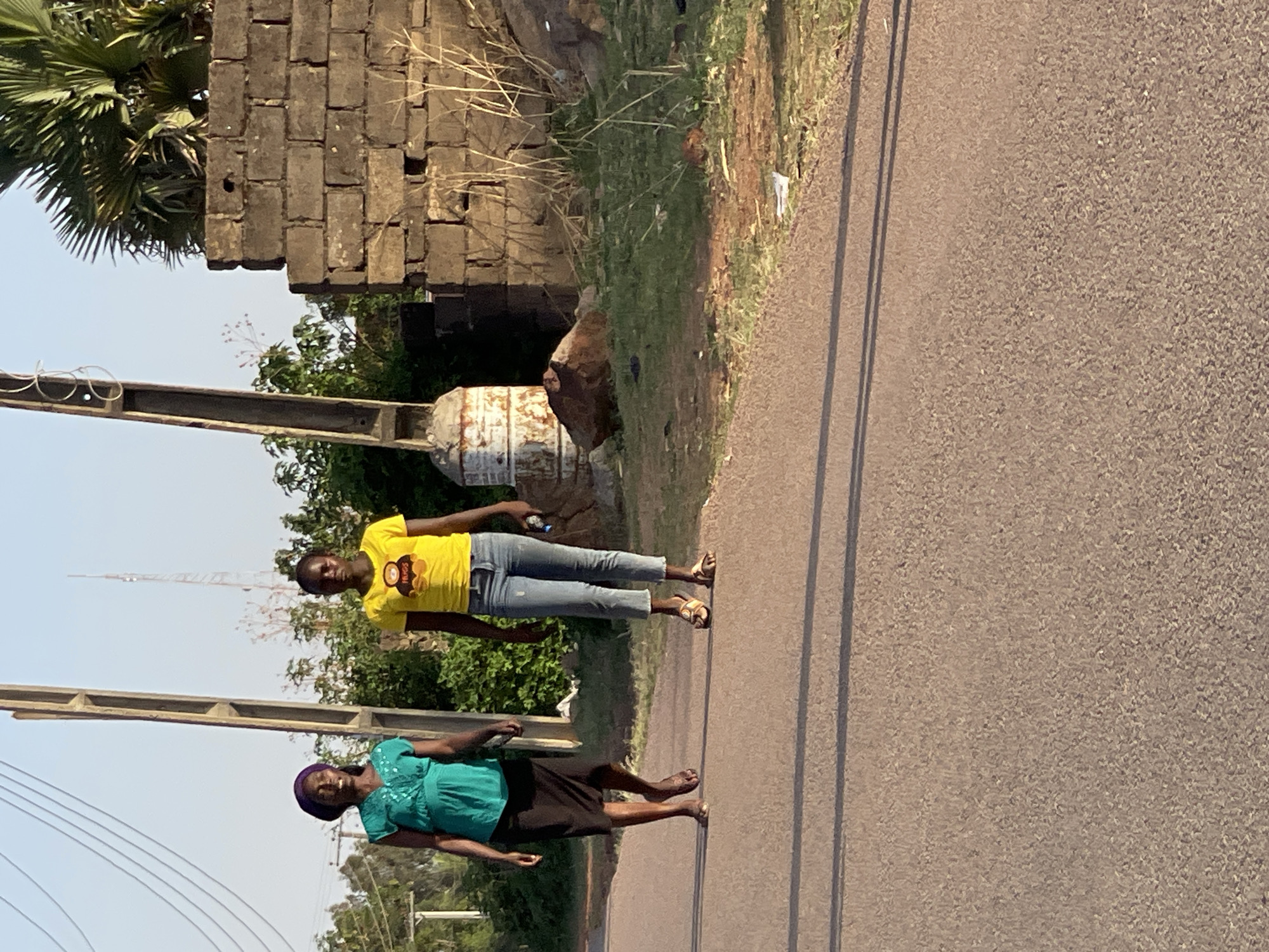 Two women are walking on a paved road in Jos, Nigeria. One woman wears a green dress and a purple head covering, and the other wears a yellow t-shirt and jeans. They are walking on the right side of the road. In the background, there is vegetation, a brick structure, and a utility pole with wires.