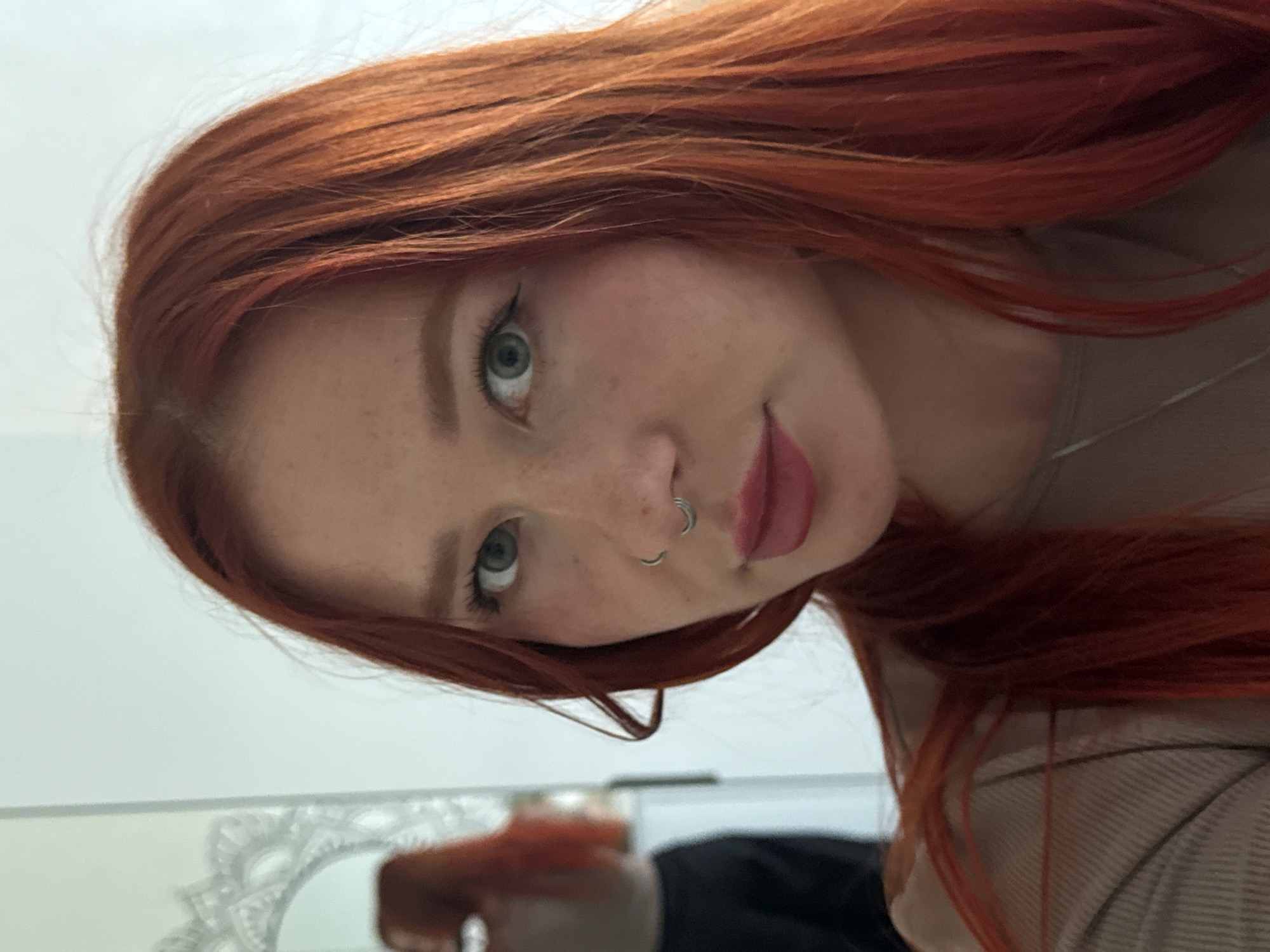 A young woman with red hair and freckles is looking at the camera. She has light green eyes and is wearing a nose ring. She is wearing a beige shirt. The background is a white wall, and a mirror is visible in the lower left corner of the frame.
The city location in Rostov-on-Don, Russia is unknown.
