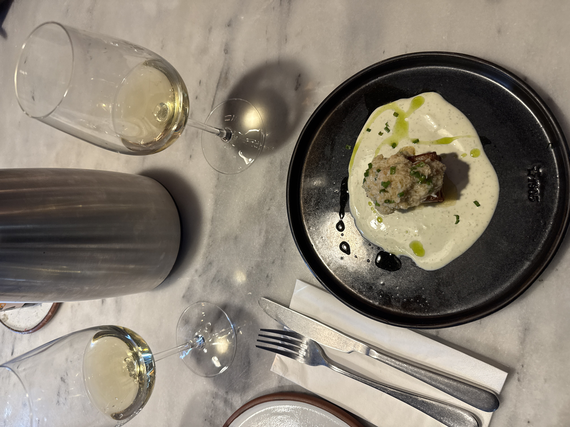The image shows a table setting on a marble surface, likely in a restaurant. There are two wine glasses, each partially filled with white wine. Beside them is a cylindrical metal object, possibly an ice bucket or a beverage container. A dark, textured plate holds a food dish: a ball of light-colored, textured substance, possibly crab or fish cake, sits atop a bed of white sauce garnished with green herbs and droplets of oil. A white napkin is placed near the plate, with a fork and knife laid on top of it. The plate bears markings or lettering that is only partially visible and may be a maker's mark. The composition is a flat lay shot from directly above, focusing on the arrangement of the food, drink, and utensils on the table. The location is not apparent and is unknown.