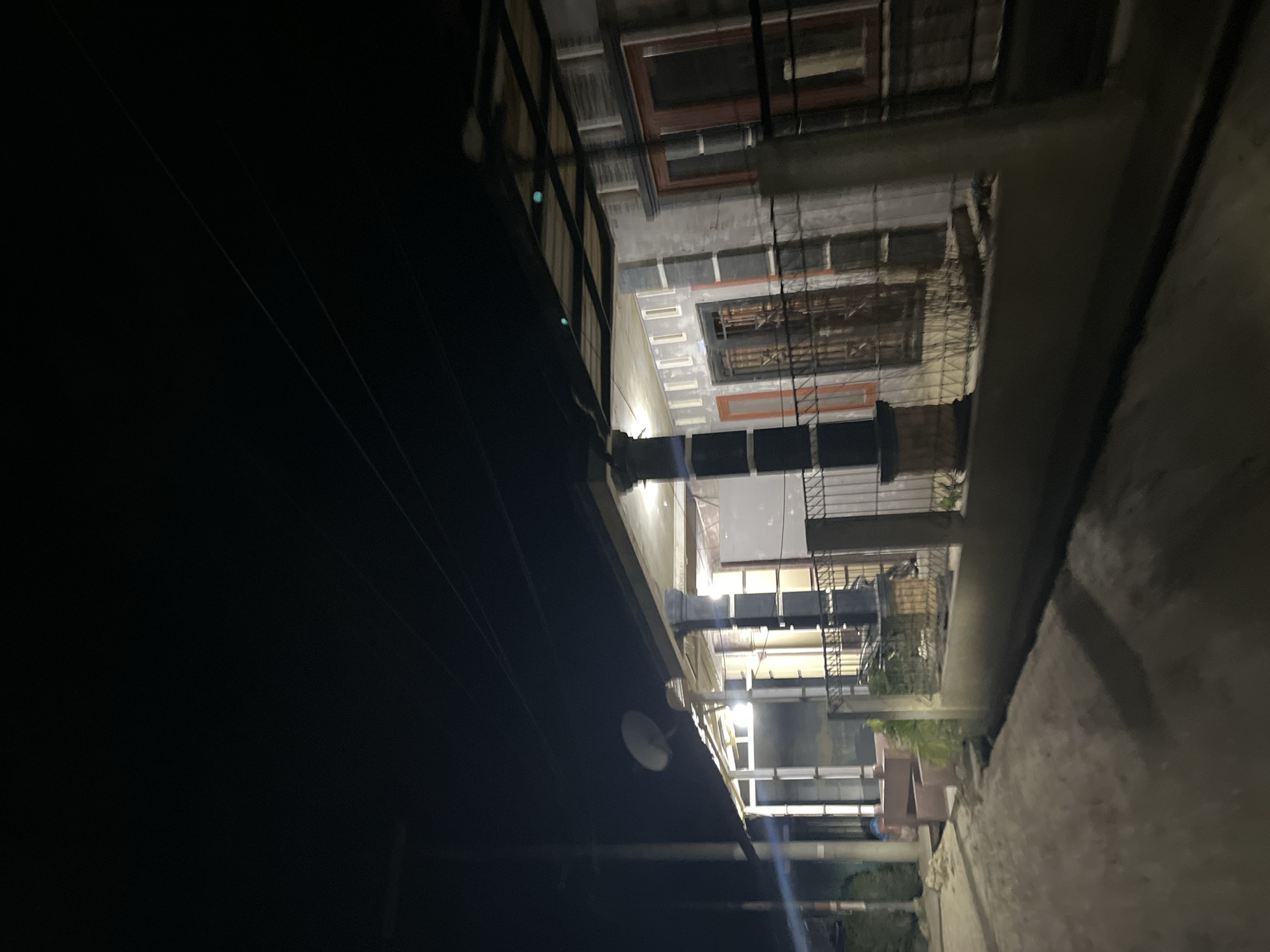 The image shows a dark, outdoor scene at night, likely in an urban or semi-urban environment in Menanggini, Indonesia. The perspective is low, looking upwards and to the right, with the camera tilted.
The primary subject appears to be a multi-story building or structure, possibly an apartment complex or a series of connected rooms. Several floors are visible, each with windows. Some windows have dark frames and appear to be made of glass, reflecting the dim light. On some floors, there are balconies with railings, and some of these balconies have what appear to be plants, suggesting an inhabited space. The architecture seems somewhat utilitarian, with concrete elements and metal railings.
In the foreground, a dark, shadowy area dominates the left side of the frame, likely a wall or a covered area, with some thin lines suggesting wires or cables running diagonally across it. A circular object, possibly a satellite dish or a light fixture, is mounted on the dark surface.
In the background, further details of the building are visible. There are multiple windows and doorways, some illuminated from within by a warm light, indicating that people are likely inside. The presence of metal bars on some windows suggests a focus on security. The scene is illuminated by artificial light sources within the building, casting shadows and highlighting architectural features.
The time of day is clearly night, and the weather appears to be calm, with no visible rain or strong winds. The location cues, such as the architecture and the mention of Menanggini, Indonesia, place the scene within that specific region. There are no people clearly visible as subjects in the image, nor any specific activity or event taking place that can be readily identified. The overall mood is somewhat subdued due to the low light conditions and the absence of clear human presence. No visible text is present in the image.