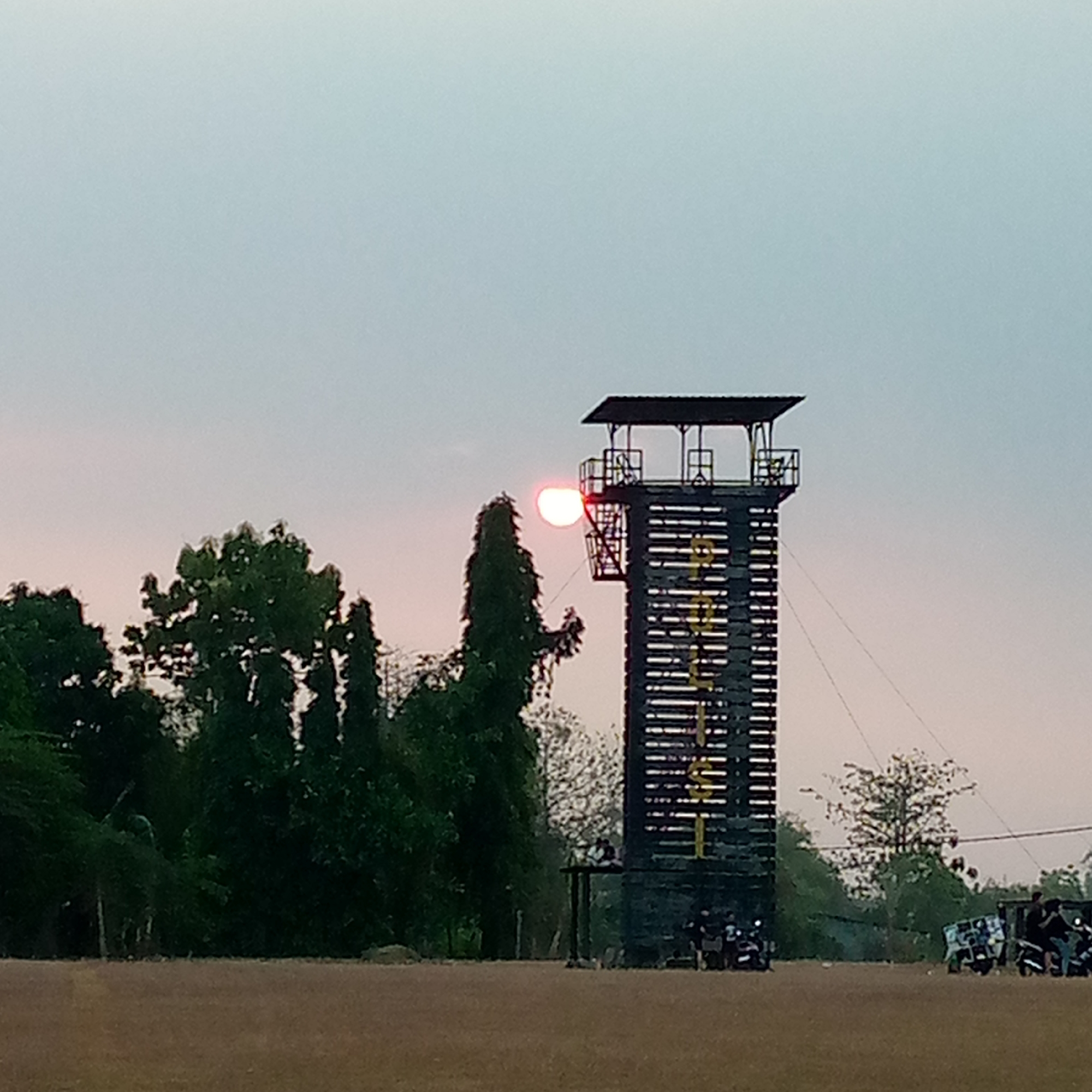 This image captures a scene at what appears to be an adventure park or training facility in Pacing, Indonesia. A tall, dark-colored tower with a viewing platform at the top stands prominently. The tower has a checkered pattern of horizontal and vertical lines, with some yellow markings visible. The sun, a bright, red-orange orb, is partially obscured by a tall, dark green tree in the foreground, positioned to the left of the tower. The sky is a pale, hazy gradient of blue and pink, suggesting it might be sunrise or sunset.
In front of the tower, several motorcycles are parked, with at least two individuals visible on or near them. A few more motorcycles and people are scattered further to the right, suggesting a gathering or event. The ground in the foreground is a flat, brownish-red surface, possibly a field or an outdoor recreational area. Lush green trees and foliage surround the area, with some taller trees on the left and more densely packed vegetation on the right. The overall atmosphere appears to be tranquil, with the muted colors and the presence of the sun low in the sky.