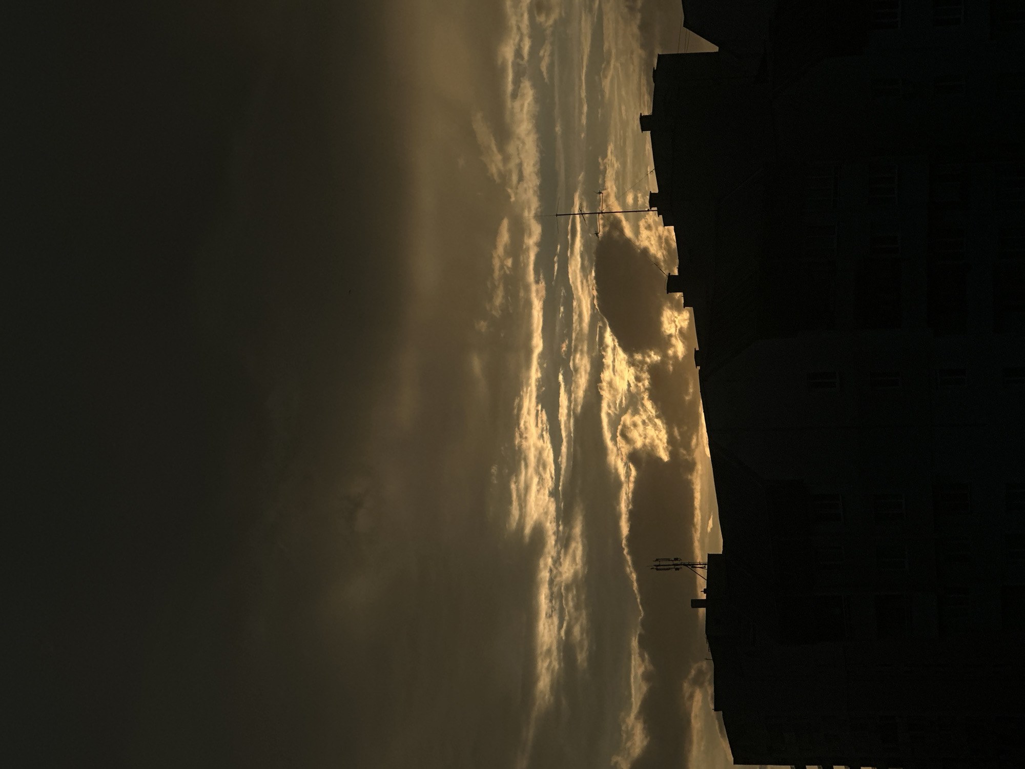 The image captures an urban scene at twilight or dawn, dominated by the dark silhouettes of multi-story buildings against a dramatically illuminated, cloudy sky.
The upper two-thirds of the frame consists of the sky, which transitions from a dark, indeterminate grey-brown at the very top to luminous golden-yellow and orange hues towards the horizon. Dense, stratified clouds partially obscure the light source, creating bright, glowing edges and streaks of light that pierce through the darker cloud formations. The varying cloud density reveals patches of intense golden light, particularly concentrated in the middle-right section of the sky.
The lower third of the image is occupied by the dark, monolithic silhouettes of several interconnected or closely positioned multi-story buildings, likely residential or mixed-use structures. These buildings are entirely underexposed, appearing as solid black forms that obscure all facade details, windows, or specific architectural features. Their rooftops present an uneven skyline, punctuated by several dark, rectangular chimney-like structures. On the rooftop of the left-most visible building, a prominent cellular antenna mast, equipped with multiple radiating elements, stands in stark silhouette against the bright sky. Towards the right, on another building, a simpler, thinner antenna pole is also visible.
No human figures, animals, or vehicles are present. No specific actions or interactions are depicted. The scene conveys a quiet, possibly desolate, urban environment during a transitional light period. The architectural style, characterized by utilitarian, multi-story apartment blocks and typical rooftop infrastructure, is consistent with residential or mixed-use areas found in post-socialist urban landscapes, including various districts within Sofia, Bulgaria. However, no unique identifying landmarks or specific features are discernible within the image to forensically pinpoint an exact address or district within Sofia.