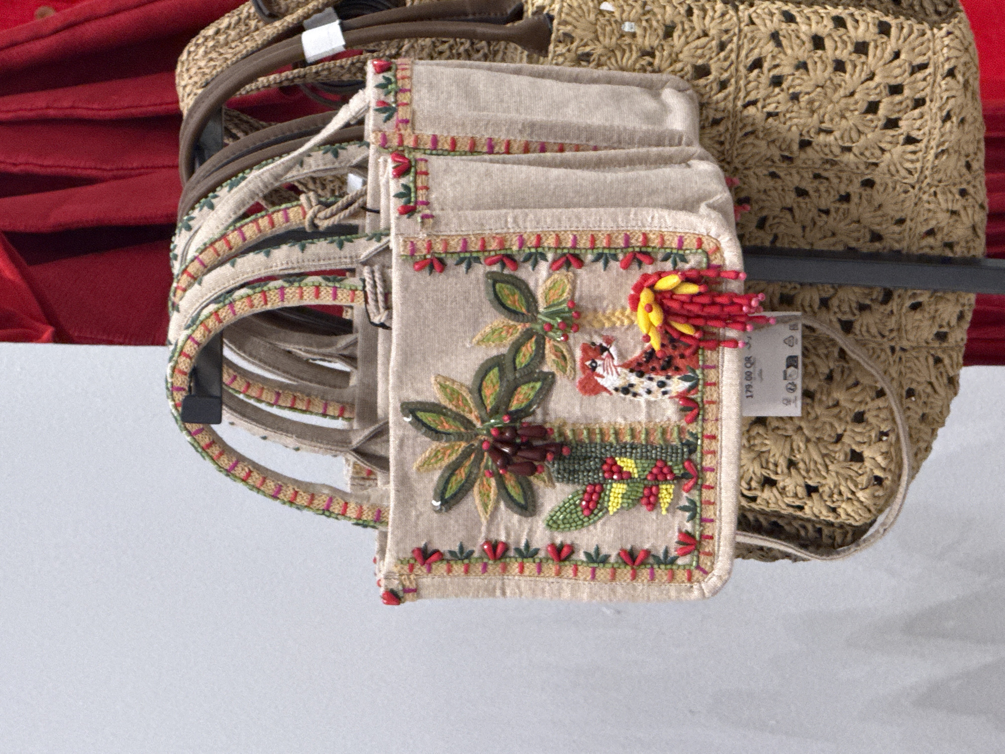 A close-up shot shows merchandise hanging from a rack in what appears to be a retail setting, potentially a shop or department store in Al Rayyan, Qatar, as inferred from the price tag showing "179.00 QR", where QR is the currency code for Qatari Riyal. A woven handbag with a cutout pattern is positioned on the right. A beige fabric wallet or small bag with embroidered floral and bird motifs is positioned in the center, overlapping the handbag. To the left are several fabric straps or belts of varying shades of beige and brown. A section of red fabric is visible in the background on the upper left side. The background beneath the hanging merchandise appears to be a light-colored wall or surface.