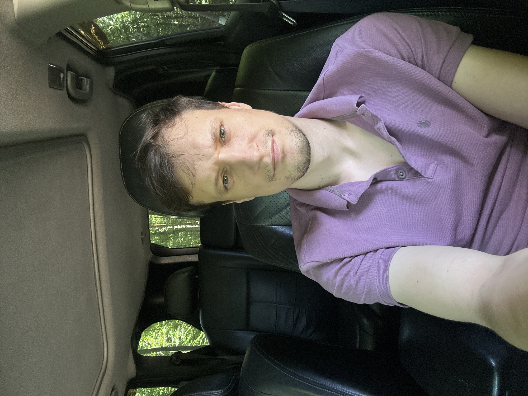 A man with light brown hair, stubble, and green eyes is sitting in the driver's seat of a car, looking at the camera. He is wearing a light purple polo shirt. The interior of the car is visible, with black leather seats and a sunroof. Trees and greenery are visible through the car windows, suggesting an outdoor, possibly rural, location.