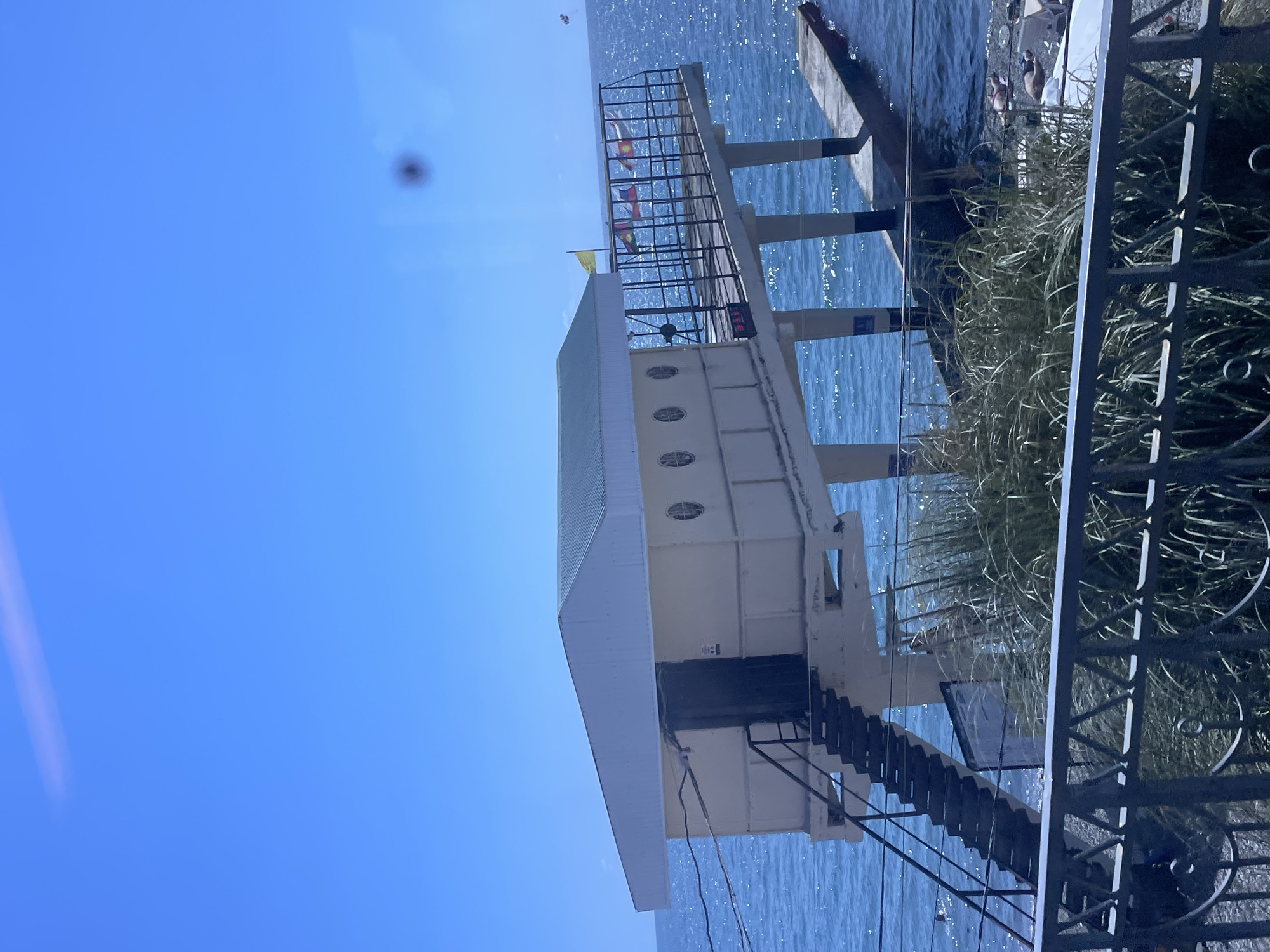 This is a daytime, outdoor, eye-level shot of a lifeguard station or similar structure situated over the water in Sochi, Russia. The building is white with a greenish roof and features several small circular windows on its side. A dark door is visible, accessed by a metal staircase. Attached to the building is a platform with a metal railing, adorned with several flags. The structure is supported by concrete pillars that extend into the blue sea. In the foreground, there is a black metal fence and tall, dry grass. The sea is calm with sunlight reflecting off its surface. In the distance, a paraglider is visible in the sky. The scene appears to be a sunny day, possibly during the summer season, with a clear blue sky.