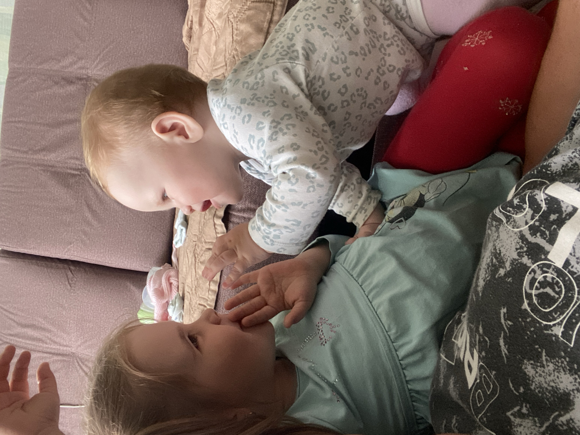 This indoor photograph, taken in Фоки, Russia, features two young children interacting on a brown or mauve upholstered sofa.
The upper child, a baby or young toddler with light blonde hair, is positioned leaning over the other child, looking down with an inquisitive expression. They are dressed in a white long-sleeved top with a light grey leopard print pattern and bright red leggings decorated with small, shiny silver snowflake designs.
Below, a slightly older toddler with blonde hair lies on their back, looking up towards the first child. This child wears a light blue long-sleeved top adorned with pink sparkly details, which appear to be an abstract design or perhaps a stylized butterfly. Their hand is raised near their mouth, possibly a gesture of curiosity or comfort.
The children appear to be engaged in a tender moment of interaction or play. A patterned fabric, likely belonging to an adult present, is partially visible on the right, featuring abstract white shapes and outlines against a grey and black background. Some shapes resemble parts of letters (e.g., "O", "D", "B", "R", "2"), though no complete words are legible. The setting is a comfortable home environment, with indirect lighting suggesting daytime. The overall impression is one of gentle affection and curiosity between the two youngsters.