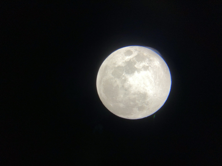 A clear, magnified image of the Moon is centered against a uniformly black night sky. The Moon appears to be in a full or near-full phase, illuminated brightly with visible surface features including numerous craters and darker maria. A subtle chromatic aberration, manifesting as a faint green and blue fringe, is discernible along the lower-left edge of the lunar disc. No other celestial bodies, man-made objects, or individuals are depicted within the frame. The observation point for this image is Buenos Aires, Argentina.