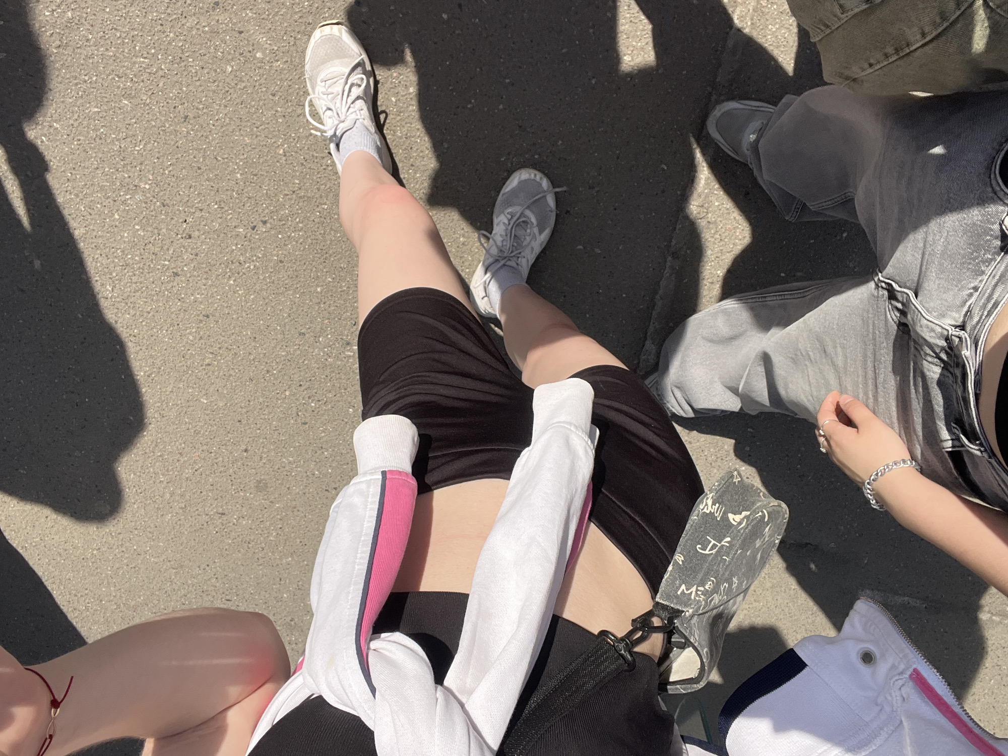 A high-angle shot depicts three individuals seated on pavement in Saint Petersburg, Russia. Sunlight casts sharp shadows across the scene.
The central figure wears black biker shorts, a white zippered jacket tied around their waist with pink accents, and white sneakers. A patterned gray purse rests on the pavement near their waist.
To the right, a person wears gray denim jeans. Their hand is visible, adorned with a silver bracelet and rings.
To the left, the edge of another person's bare thigh is visible, featuring a red ankle bracelet.
The pavement appears to be asphalt.