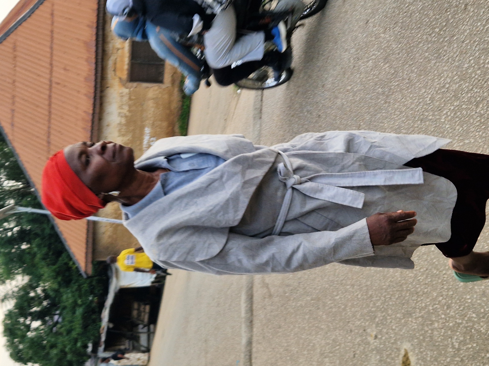 The image shows an outdoor scene in Jos, Nigeria. In the foreground, a woman wearing a red headscarf, a light grey open coat, and dark trousers walks towards the left of the frame. She has a grey garment underneath her coat and is wearing light green footwear. To the right of the woman, a person rides a motorcycle, wearing a blue helmet and a grey jacket. The background features a building with an orange tiled roof and a yellow-shirted person standing near a structure. The environment appears to be a street or pathway with a rough, textured surface.