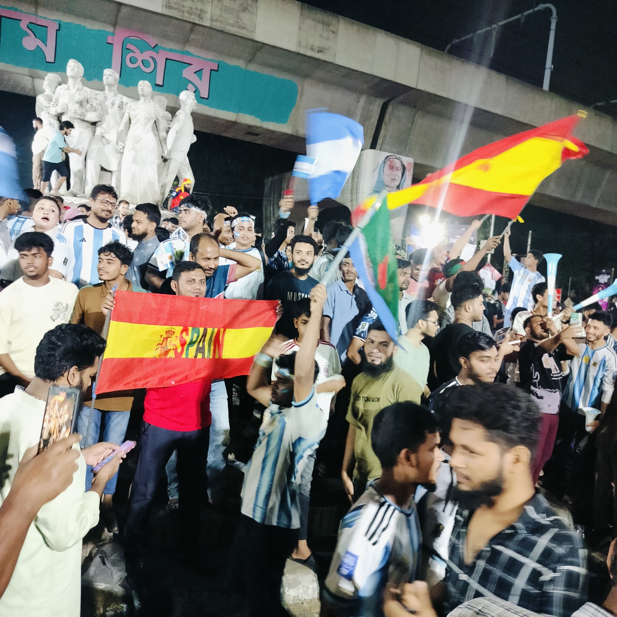 In Dhaka, Bangladesh, a crowd gathers at night, possibly celebrating a sporting event. Several individuals are seen holding flags, including one depicting the colors of Spain and the word "SPAIN" emblazoned on it, and another with the blue and white stripes characteristic of Argentina. Some individuals are wearing face paint or headbands in these team colors. In the background, a large mural with Bengali script and statues of people are visible. The scene is illuminated by artificial light, creating a festive atmosphere among the onlookers.