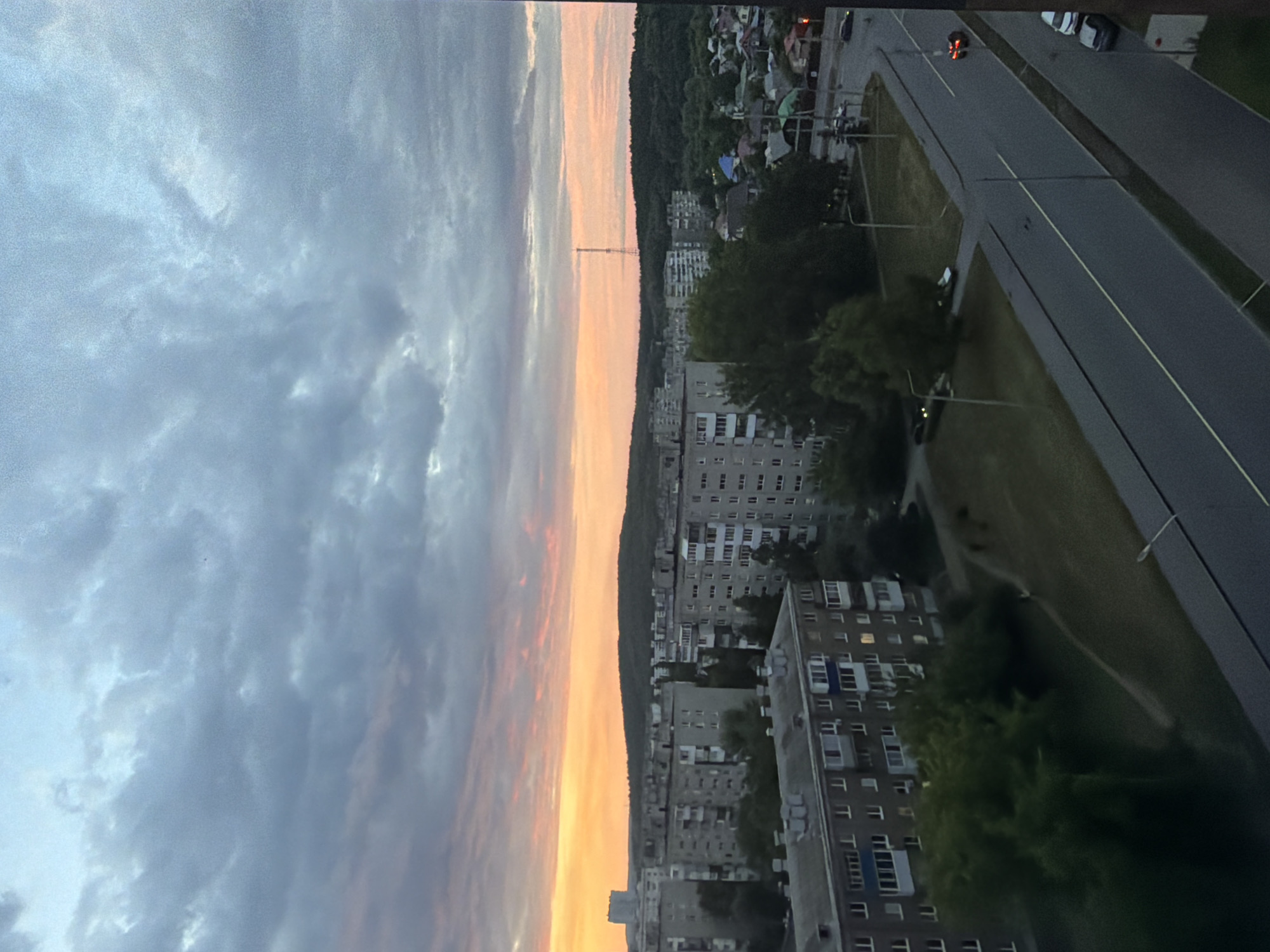 The image presents an expansive urban landscape viewed from a high vantage point, likely during dusk or dawn. The upper portion of the frame is dominated by a dramatic sky, featuring a transition from dark grey and blue clouds at the top to vibrant orange, pink, and yellow hues along the horizon, characteristic of sunrise or sunset.
Below the colorful sky, a large, dark, tree-covered hill or mountain range stretches horizontally across the midground. A tall, slender communication tower is silhouetted against the bright horizon, positioned on or just behind this hill.
The foreground and midground reveal a residential area with numerous multi-story apartment buildings, featuring rows of rectangular windows and balconies. Some windows within these buildings show illuminated lights. A multi-lane asphalt road traverses the lower right and foreground, marked by a yellow dividing line. Several vehicles are present: a few cars are parked, and a red vehicle with illuminated tail lights is seen moving on the road. Green spaces, including grass and trees, are interspersed among the buildings and alongside the road, with a narrow dirt path visible in one grassy section. The architectural style of the buildings suggests a possible Eastern European or post-Soviet urban environment. No discernible text is visible.