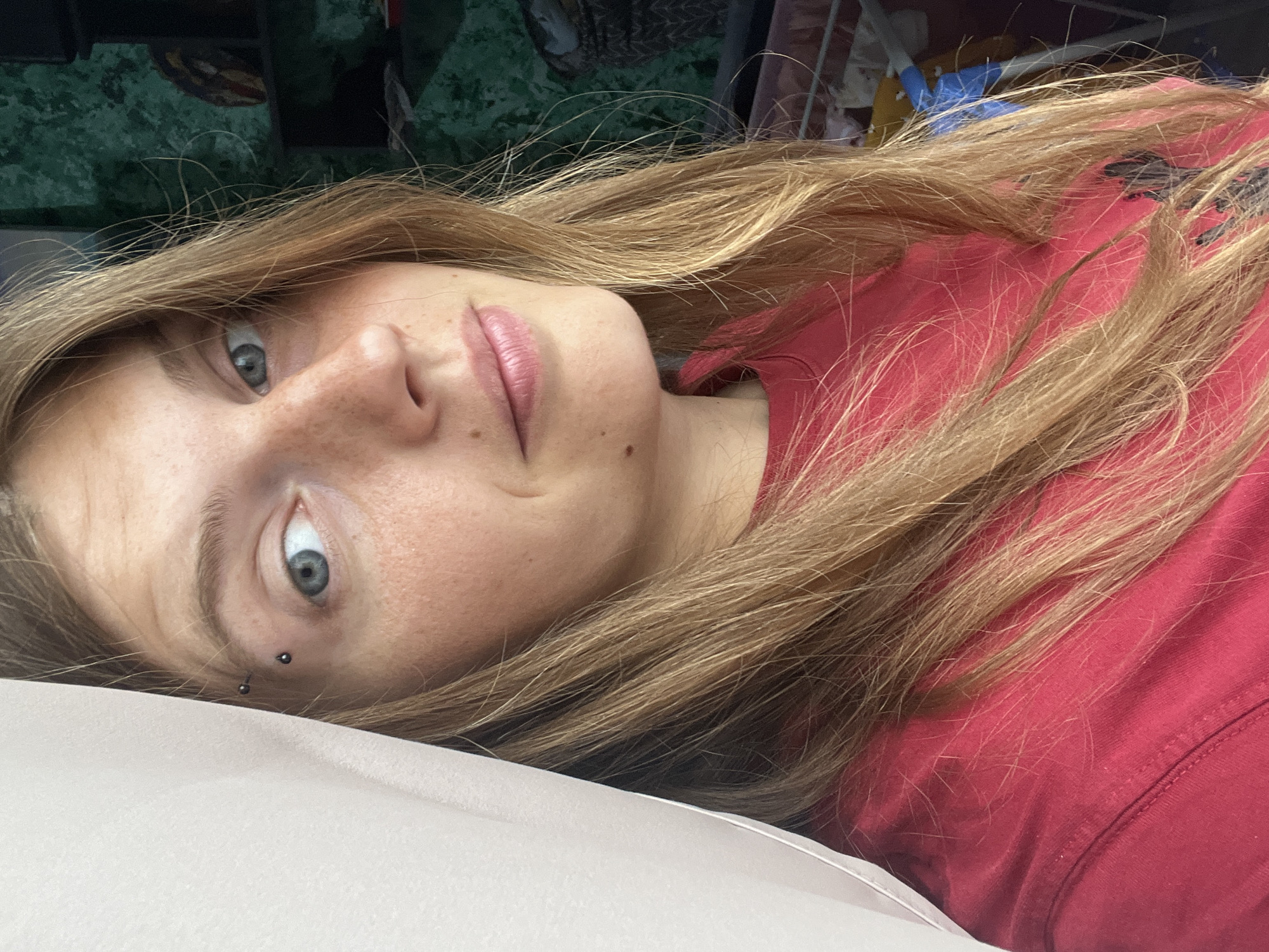 This is a close-up, indoor selfie of a woman with long, blonde hair and blue eyes. She is wearing a red t-shirt. She has freckles on her face and an eyebrow piercing. The background appears to be a wall with a green, textured pattern, and some shelves with objects on them. A white curtain is visible on the left side of the image. The lighting suggests it is daytime. The image does not provide any specific indicators of location within the city of Сеніцкі сельскі Савет, Belarus, or any particular urban setting.