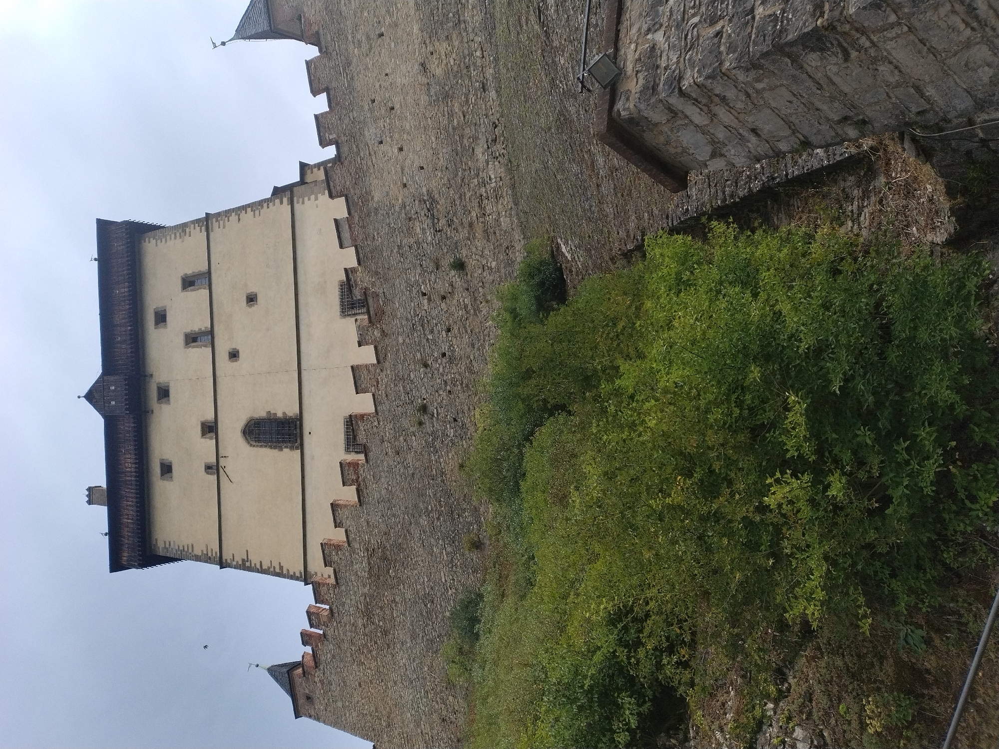 An upward-angle shot captures a imposing medieval castle against a cloudy sky. The castle's facade is a pale yellow, accented with darker stone and brickwork, featuring several small rectangular windows and a larger arched window with a lattice. The castle walls are crenelated with a rough stone lower section and a lighter stone upper section, topped with a dark wooden roof structure. To the left, a dense, vibrant green bush grows on a rocky slope, obscuring the base of the castle wall. Further down, a stone wall section with a light fixture is visible. The scene is located at Karlštejn Castle in Karlštejn, Czechia.